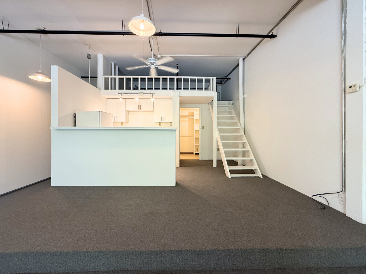 A spacious, open-concept living area features a white kitchen with modern cabinetry and a refrigerator. The kitchen island serves as a divider from the main space. A staircase leads to a loft area above, while the floor is covered in gray carpeting. Soft lighting fixtures hang from the ceiling, and a ceiling fan is visible. The walls are painted white, creating a bright and airy atmosphere.