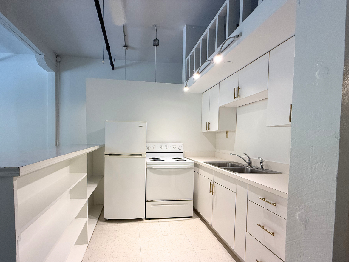 A compact kitchen featuring white cabinetry and appliances. The kitchen includes a refrigerator, an electric stove with an oven, and a double sink. The countertops are light-colored, and there are gold handles on the cabinets. A shelf runs along one side, providing additional storage space. The walls are painted in a light color, contributing to a bright and airy atmosphere.