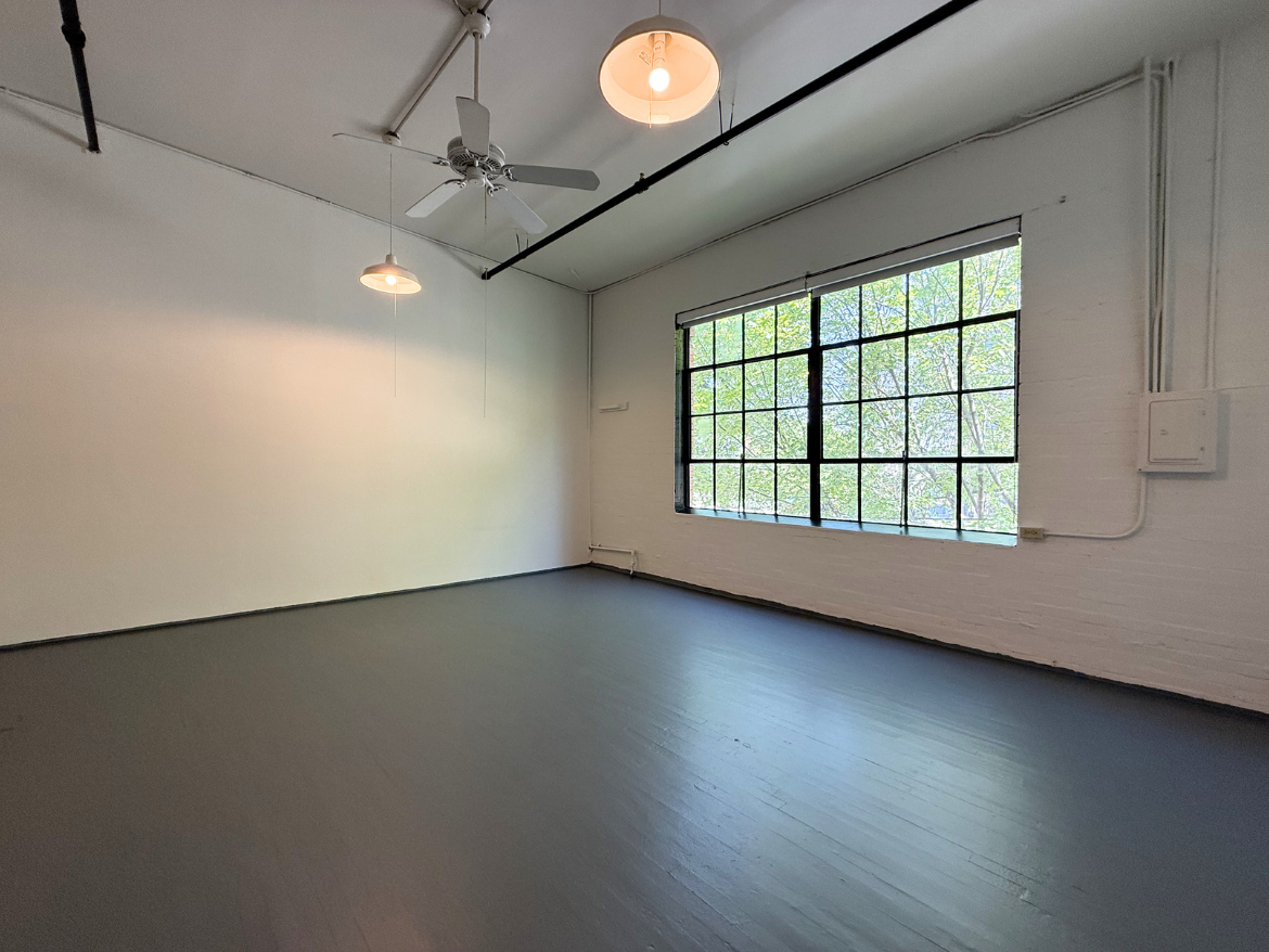 A spacious, empty room features a polished gray floor and white brick walls. Two pendant lights hang from the ceiling, providing soft illumination. A large window with multiple panes allows natural light to fill the space, showcasing greenery outside. The overall atmosphere is bright and airy, ideal for various uses.