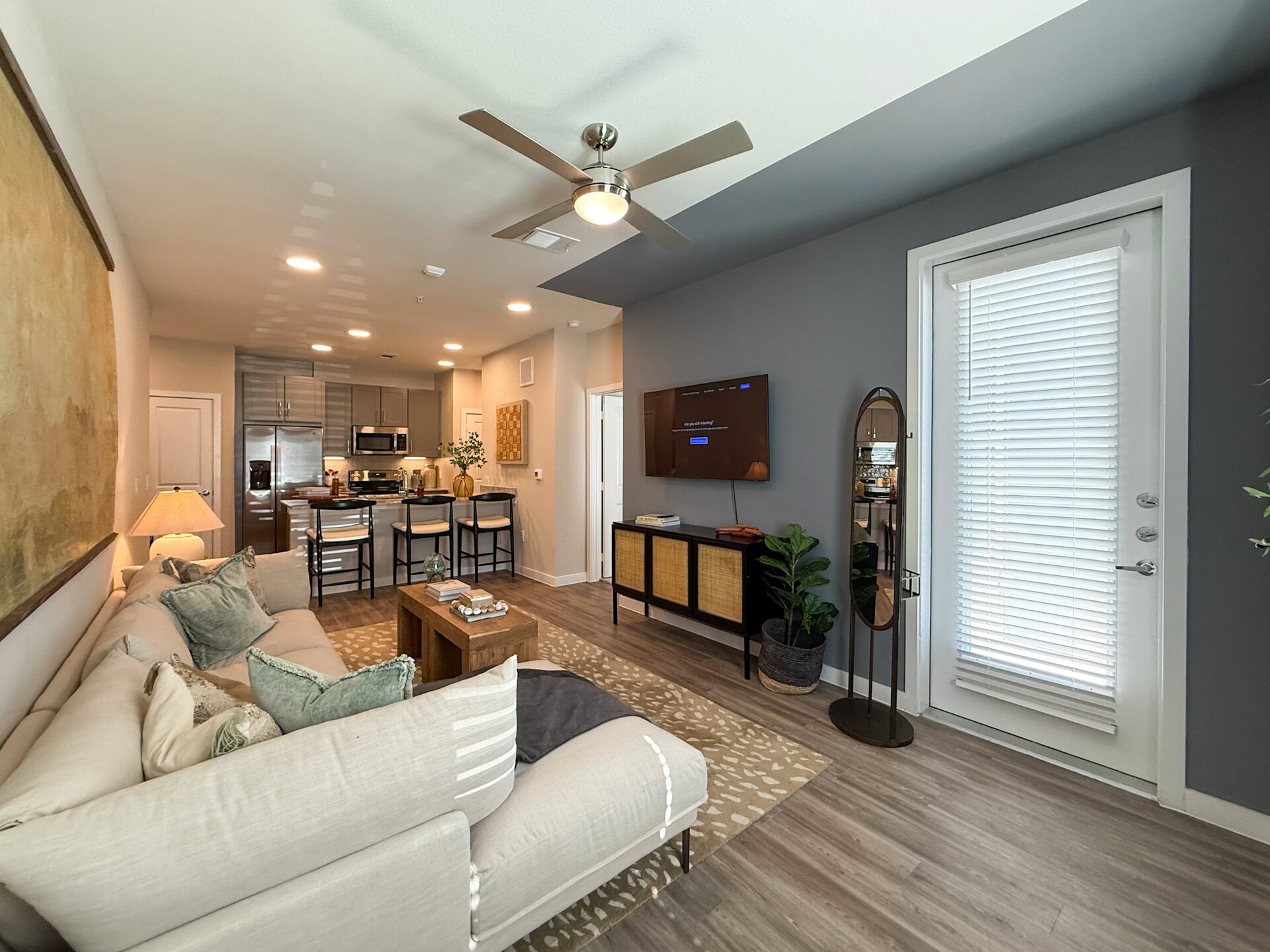 A modern San Antonio apartment living room features a spacious living space. In the background, a kitchen area showcases stainless steel appliances and bar stools. Natural light filters through a door with blinds, and a tall mirror stands beside a potted plant, enhancing the room's contemporary aesthetic.
