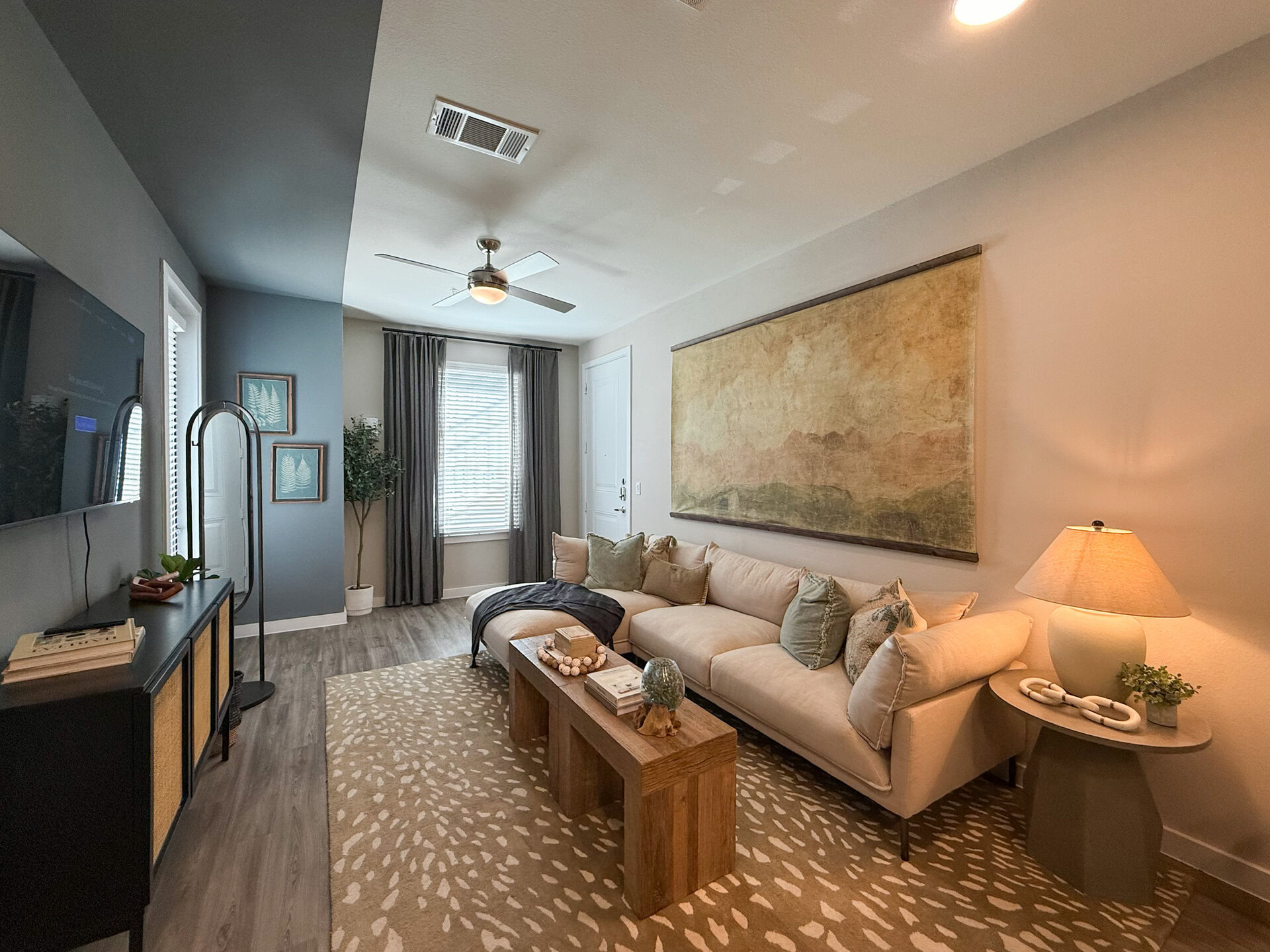 A cozy San Antonio apartment living room features a spacious open layout. The room is illuminated by natural light streaming through the window, which is dressed with gray curtains. The flooring is a warm wood finish, complemented by a patterned area rug.
