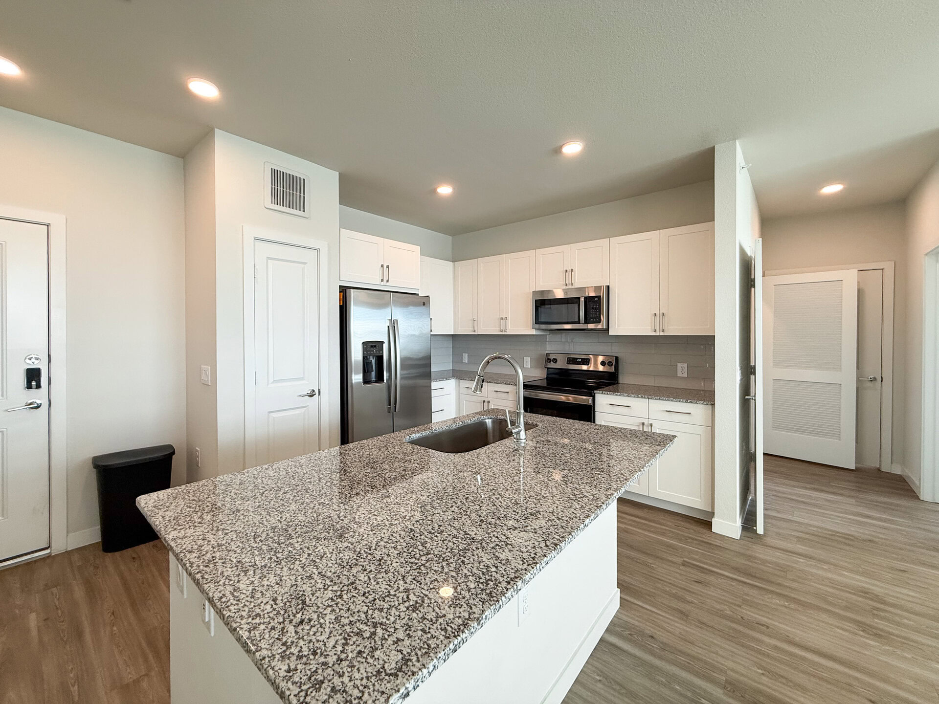 A modern San Antonio apartment kitchen featuring white cabinetry and a large granite island with a sink. Stainless steel appliances, including a refrigerator, microwave, and oven, are integrated into the design. The backsplash is a light gray tile, complementing the overall aesthetic. The space is well-lit with recessed lighting.