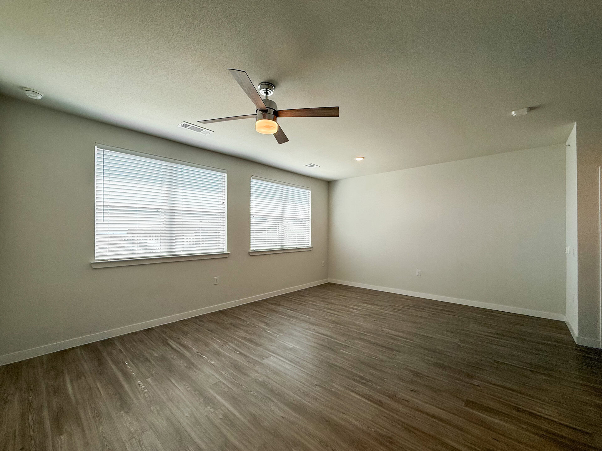 A spacious San Antonio apartment living room with light-colored walls and a wood-like floor. Two large windows with white blinds allow natural light to fill the space.