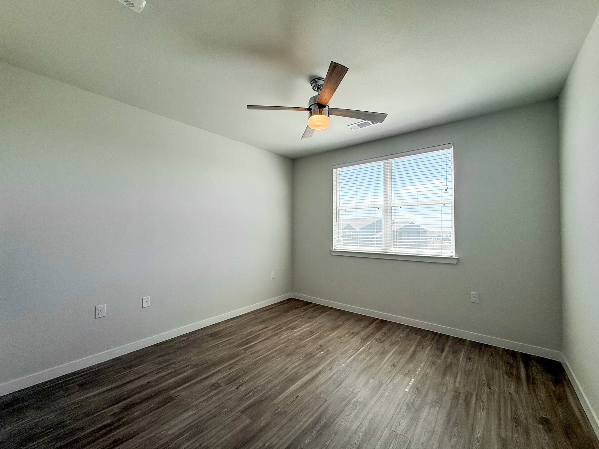 A bright, San Antonio apartment bedroom features light-colored walls and a wooden floor. Natural light streams in through a window with white blinds.