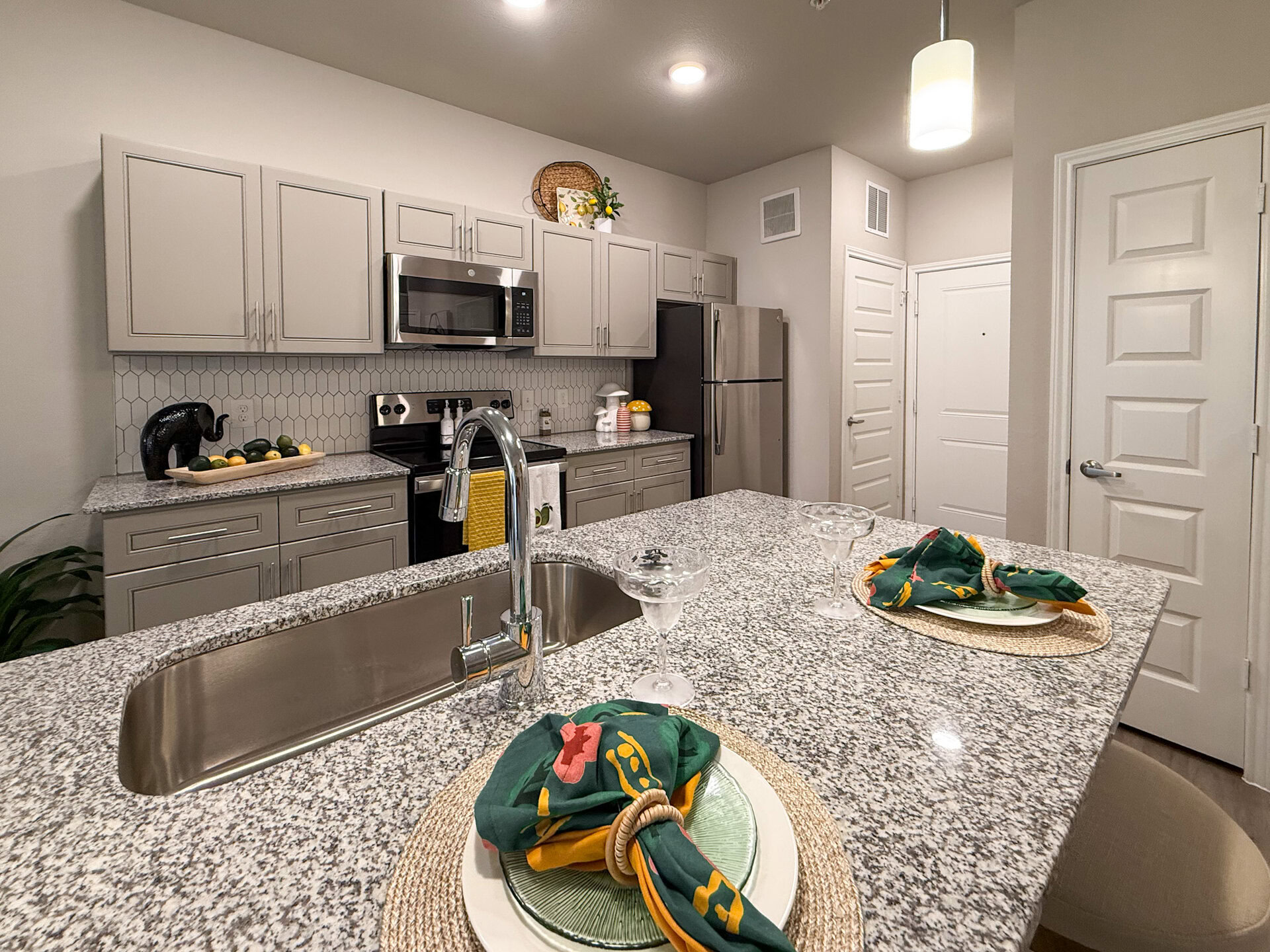A modern San Antonio apartment kitchen featuring light gray cabinetry and a granite countertop. The kitchen is equipped with stainless steel appliances, including a microwave and refrigerator. The backsplash consists of white hexagonal tiles.