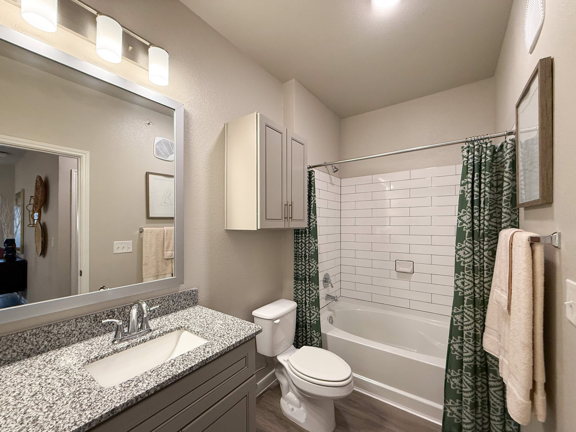 A modern San Antonio apartment bathroom features a light-colored wall with a large mirror above a granite countertop sink. The sink has a sleek faucet. To the right, a bathtub is enclosed with white subway tiles and a green patterned shower curtain. Above the sink, there are two cabinets for storage. A towel rack holds a beige towel. The overall design is clean and contemporary.