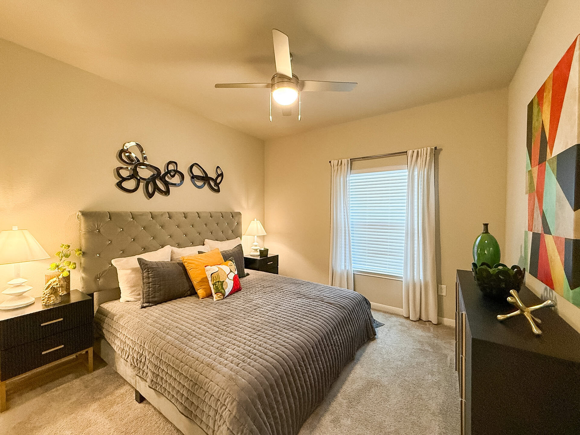 A cozy San Antonio apartment bedroom features a spacious layout. The room is illuminated by a ceiling fan light and has a window with white blinds, framed by sheer curtains. The carpeted floor enhances the room's warmth and comfort.