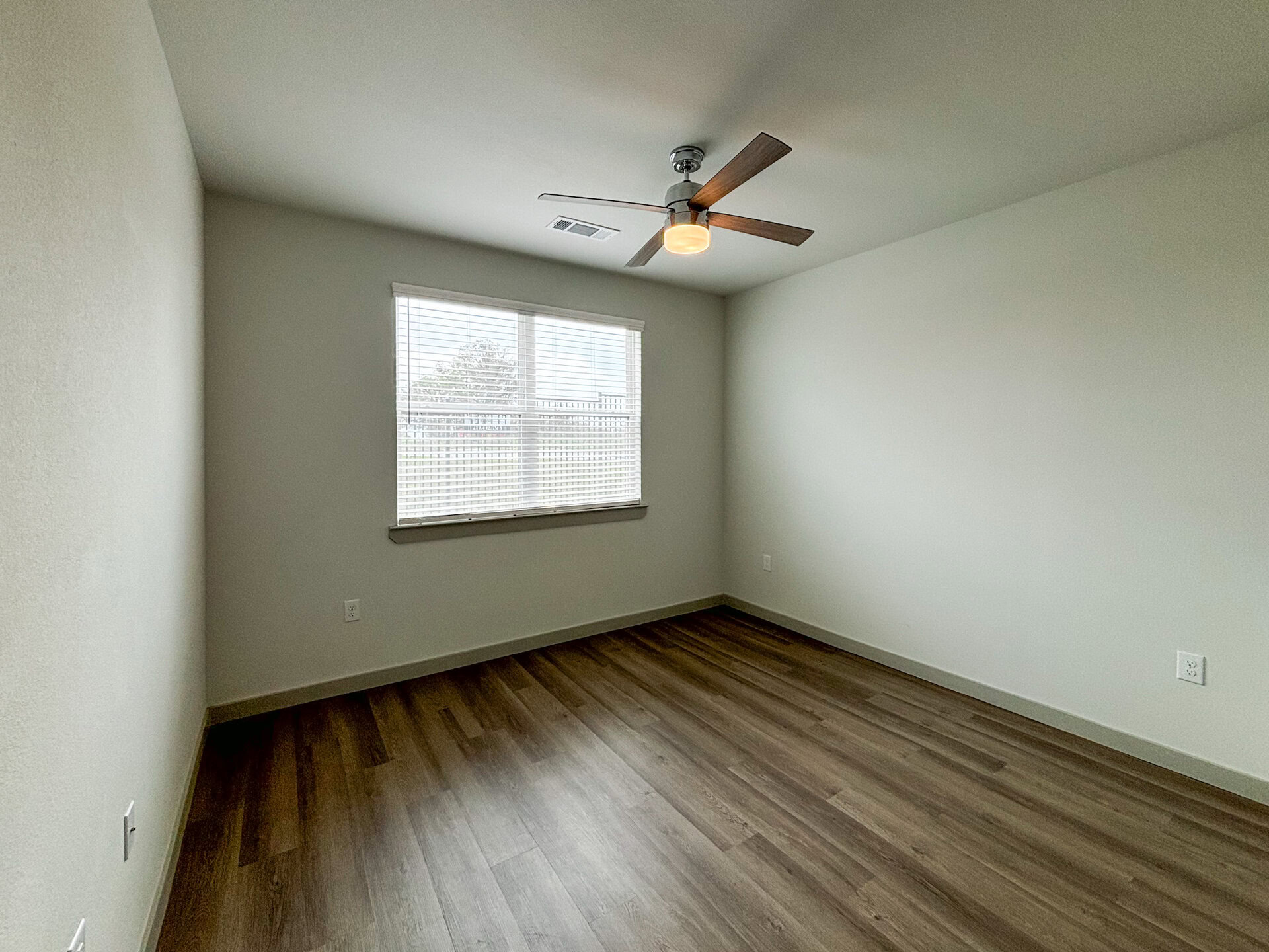 A Northwest San Antonio spacious bedroom features light-colored walls and a wooden floor. A ceiling fan with wooden blades is mounted in the center, and a window with blinds allows natural light to enter.