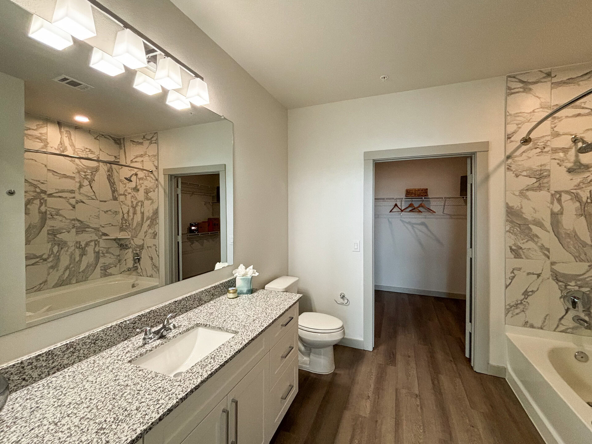 A modern San Antonio apartment bathroom features a large mirror above a granite countertop with a single sink. To the right, a white toilet is positioned next to a doorway leading to a closet with hanging space. The walls are decorated with large marble-patterned tiles, and a shower is visible in the corner. Bright, square light fixtures illuminate the space, which has a warm wood-like floor.