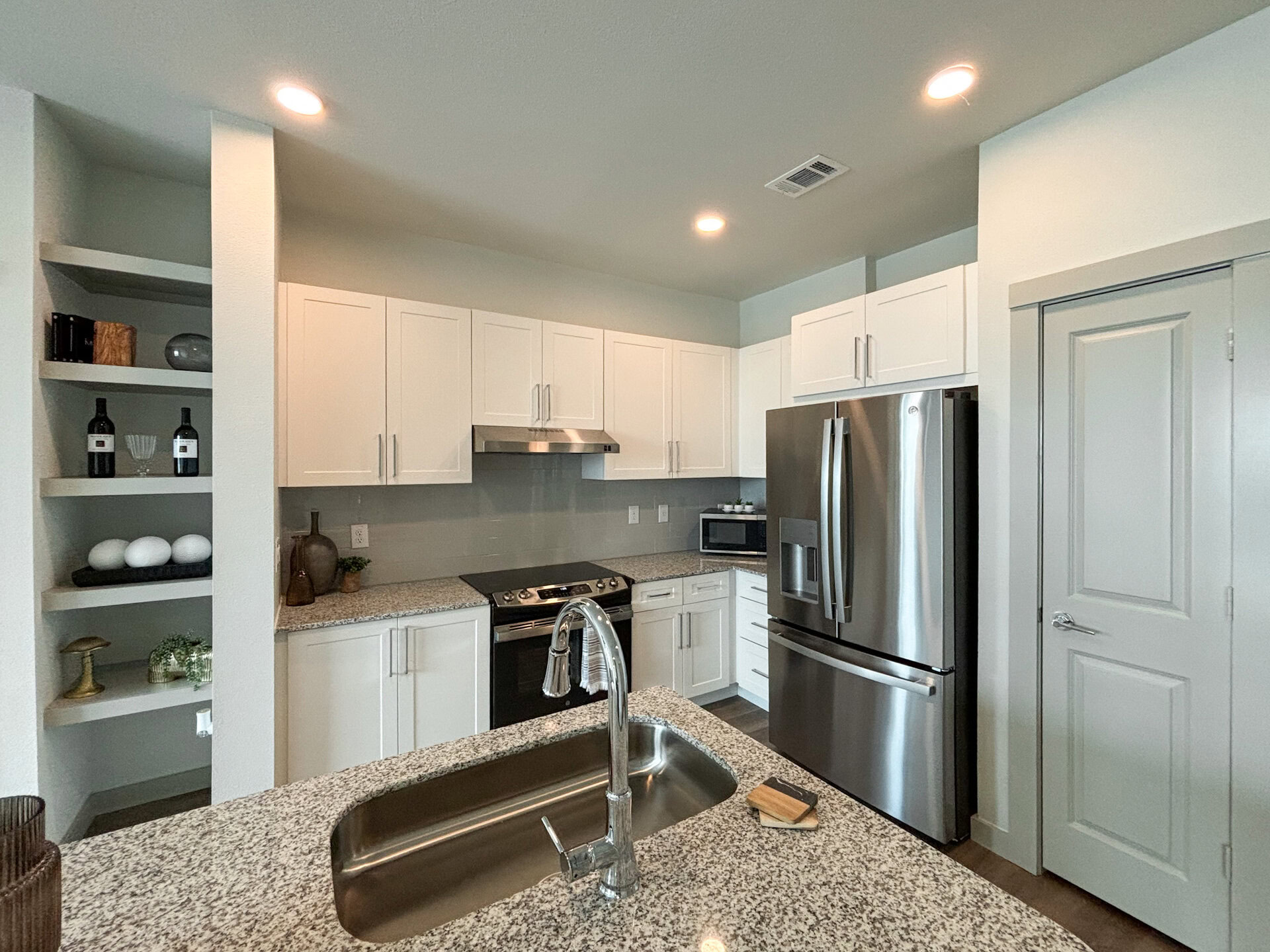 A modern San Antonio apartment kitchen featuring white cabinetry with sleek handles, a stainless steel refrigerator, and a black stove with a stainless steel hood. The countertops are made of speckled granite, and there is a stainless steel sink with a pull-down faucet. Open shelving on the left. The walls are painted in a light color, contributing to a bright and airy atmosphere.