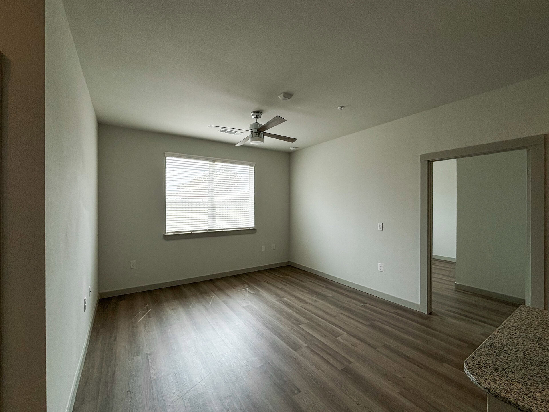 A spacious San Antonio apartment living room features light gray walls and a ceiling fan with wooden blades. Natural light streams in through a window with horizontal blinds, illuminating the light-colored flooring. The overall atmosphere is bright and minimalistic.