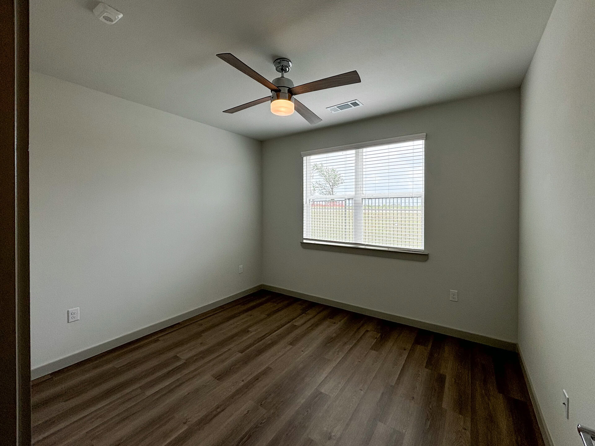 A San Antonio apartment bedroom, well-lit room features a ceiling fan with wooden blades and a light fixture. The walls are painted a light color, and the floor is covered with laminate wood. A large window with white blinds allows natural light to enter.