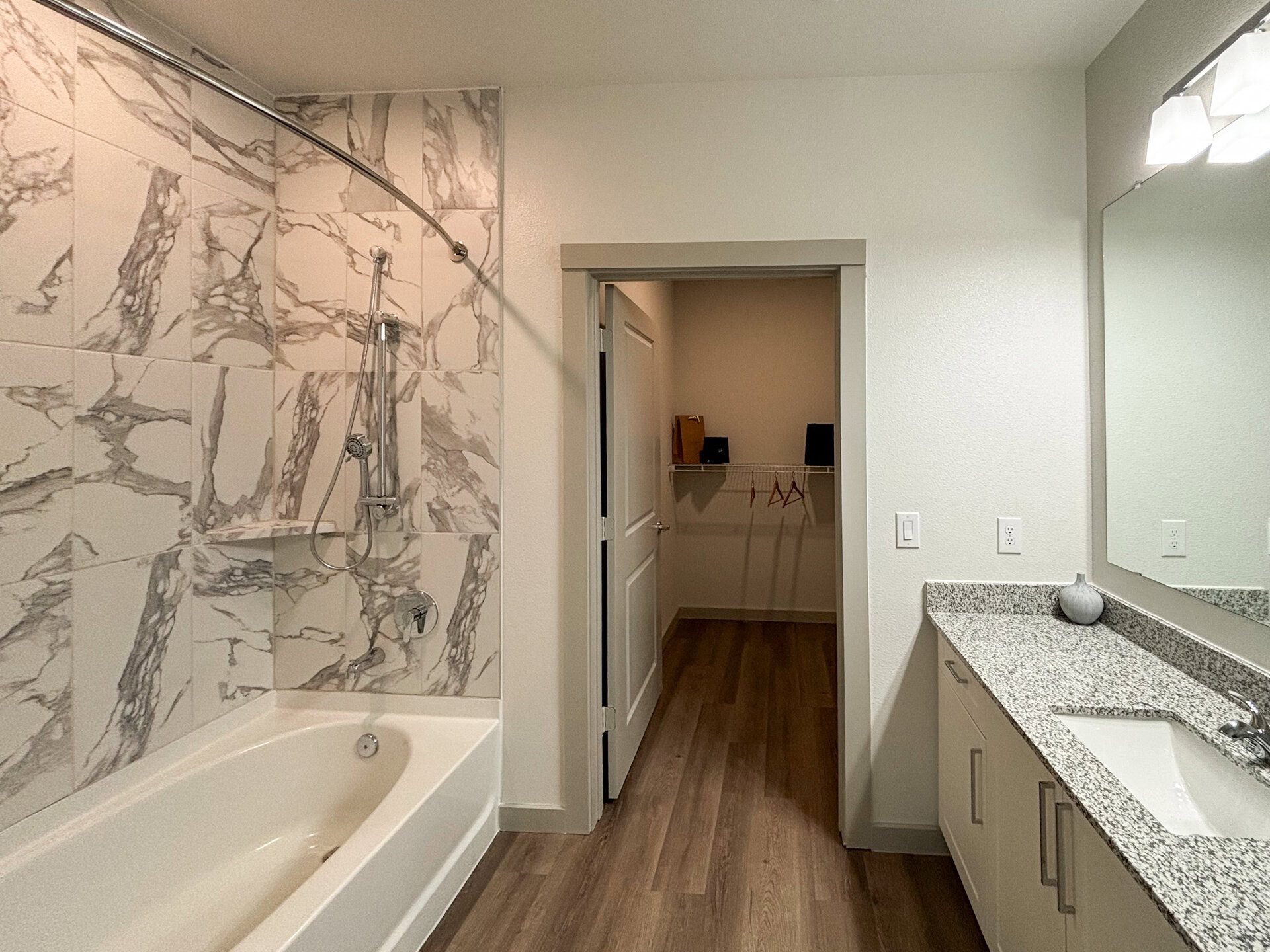 A modern San Antonio apartment bathroom features a bathtub with a showerhead mounted on a curved rod above. The walls are adorned with large, marble-patterned tiles in shades of white and gray. To the right, a vanity with a granite countertop includes a sink. A mirror hangs above the vanity, reflecting the space. A doorway leads to a small closet with a shelf and hangers, while the floor is finished with light wood planks. The overall color scheme is neutral and contemporary.
