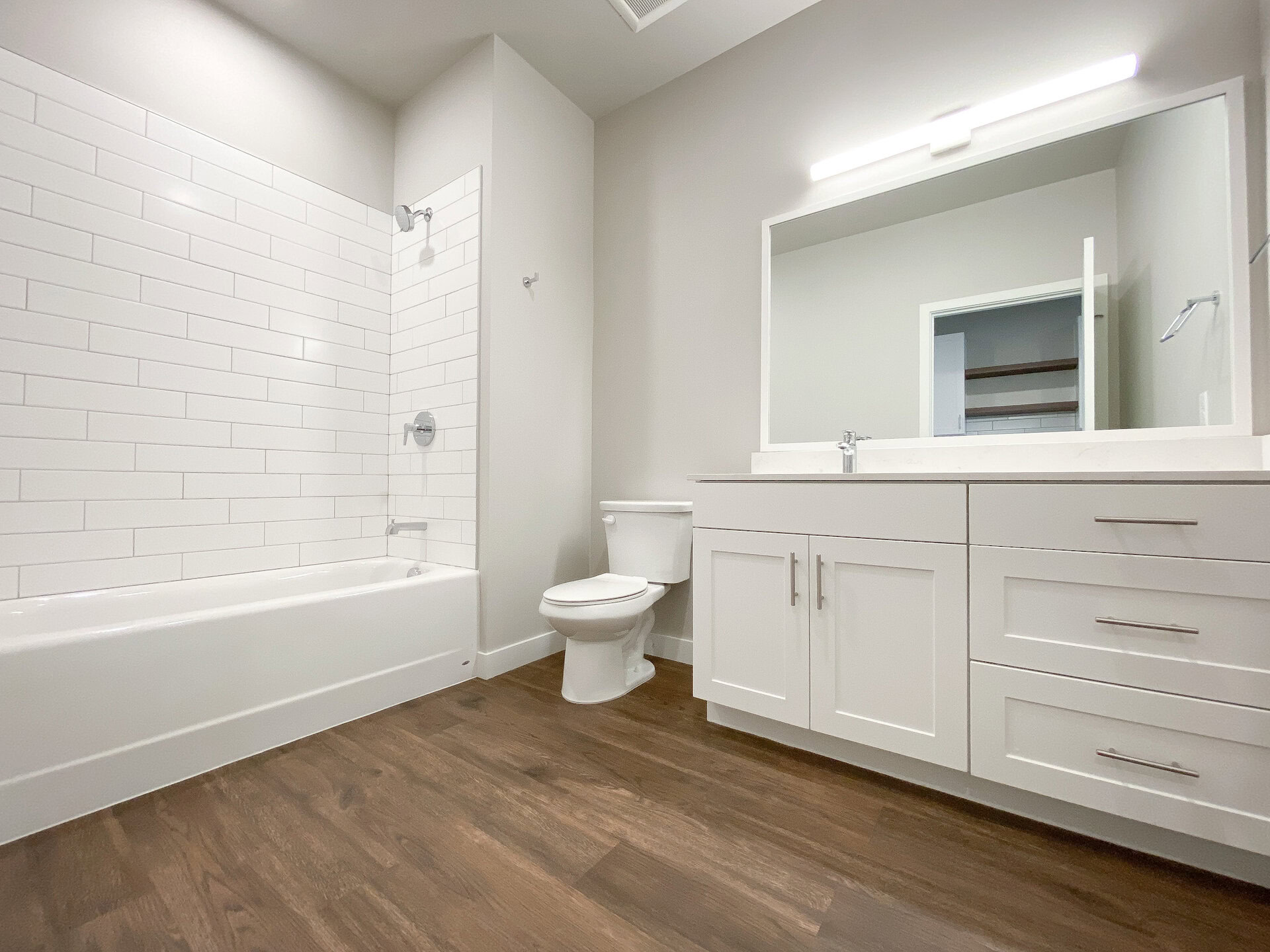 A modern bathroom features a white tiled shower area with a bathtub, complemented by a sleek chrome showerhead and faucet. The flooring is a warm wood tone, contrasting with the white cabinetry. A white vanity with a marble countertop and a large mirror above provides ample storage with multiple drawers and a cabinet. A white toilet is positioned next to the vanity, and a small towel rack is mounted on the wall. The overall design is clean and contemporary, with neutral wall colors enhancing the bright atmosphere.