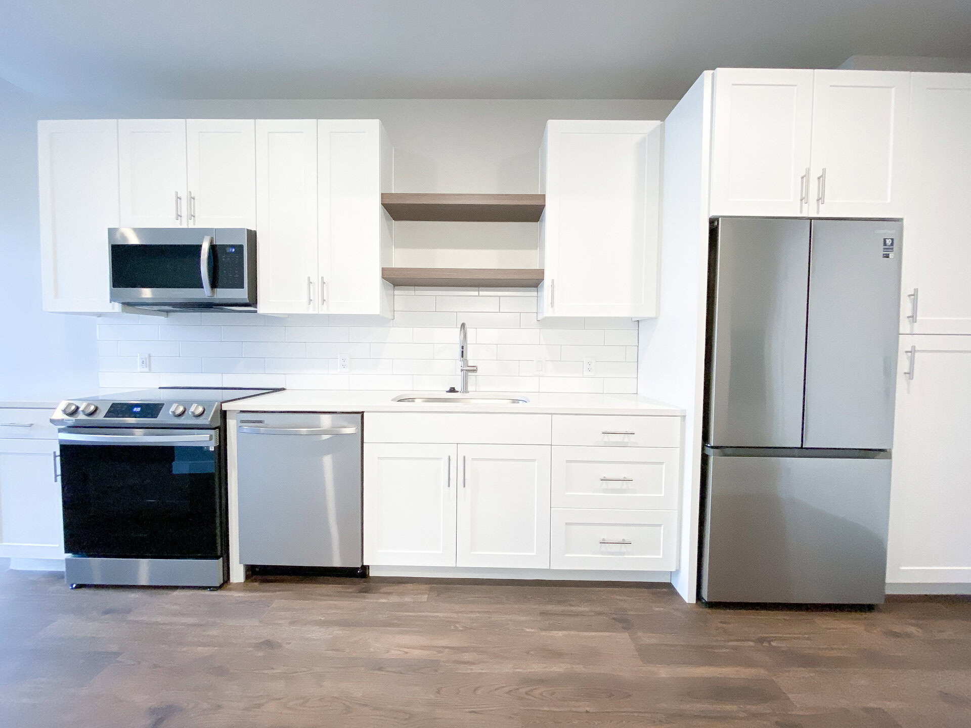 A modern kitchen features white cabinetry with sleek silver handles, a stainless steel refrigerator, and a built-in microwave above a black oven. A silver dishwasher is positioned next to the oven. The countertop is white with a stainless steel sink and a contemporary faucet. The backsplash consists of white subway tiles, and there are open wooden shelves above the sink for additional storage. The flooring is a warm wood tone, enhancing the kitchen's contemporary aesthetic.