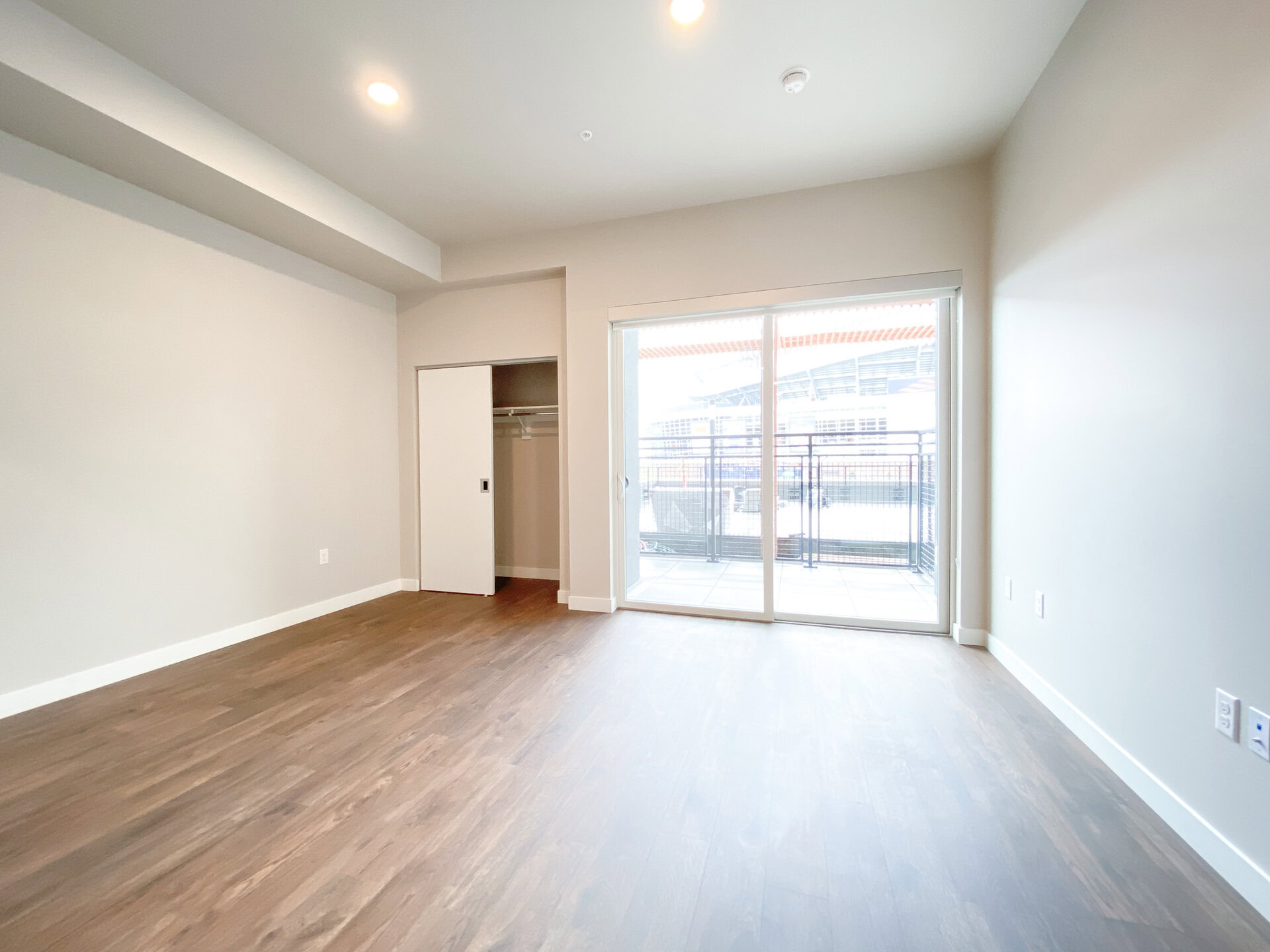 A spacious room features light-colored walls and a large window with sliding glass doors that open to a balcony. The floor is made of wood, and there is a small closet with sliding doors on the left side. The overall ambiance is bright and modern, with ample natural light coming in from the balcony.