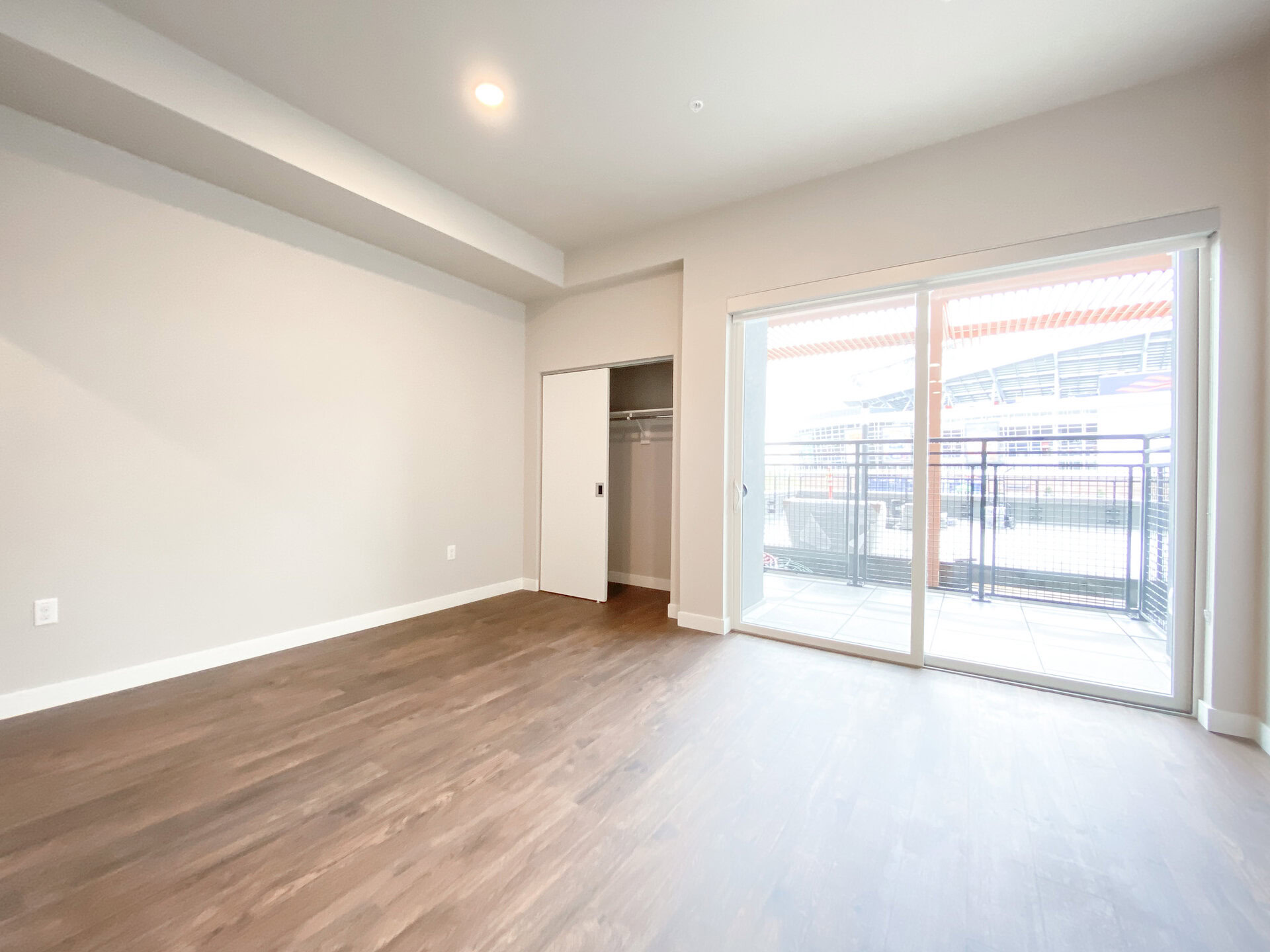 A spacious room features light-colored walls and a wooden floor. On one side, there is a closet with sliding white doors, partially open to reveal a hanging rod. Large glass doors lead to a balcony with a view of an outdoor area, allowing natural light to fill the space. The overall ambiance is modern and inviting.