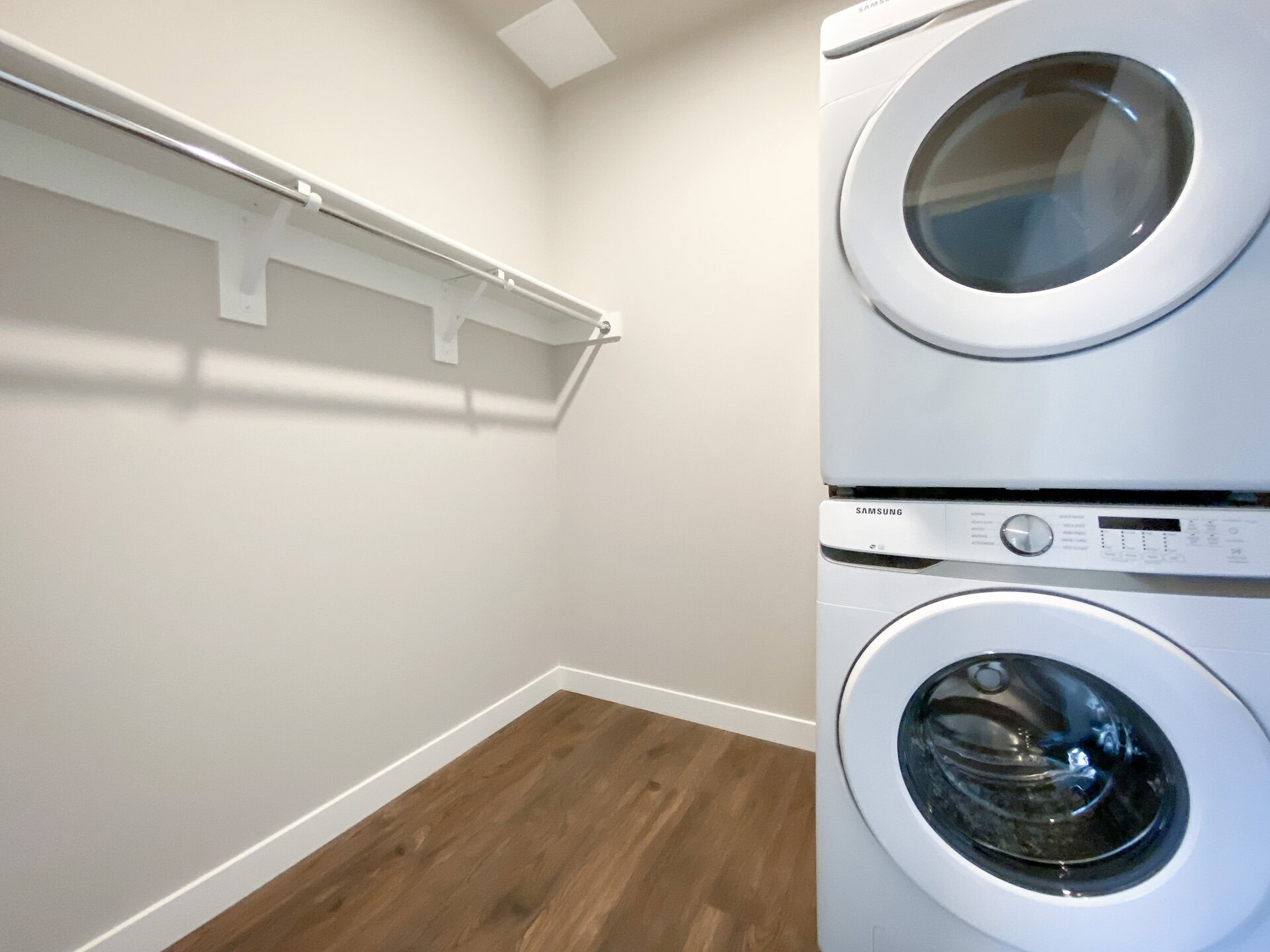 A compact laundry room features a stacked washer and dryer in white, positioned against a light-colored wall. Above the appliances, there is a metal rod mounted on brackets, likely for hanging clothes. The flooring is a warm wood tone, adding a cozy feel to the space. The overall design is minimalistic and functional.