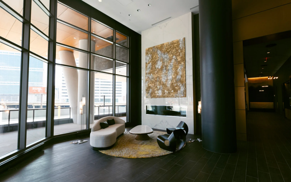 A modern lobby features large glass windows allowing natural light to flood the space. In the center, a curved white sofa with black cushions is paired with a sleek black chair. A round coffee table sits atop a textured yellow and gray area rug. Behind them, a striking gold and white artwork is mounted on a marble wall, complemented by a contemporary fireplace below. The flooring is dark wood, enhancing the elegant atmosphere.
