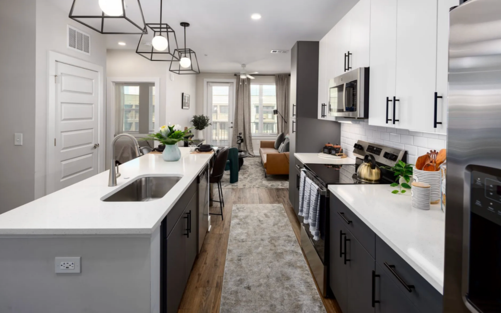 A modern kitchen features a central island with a white countertop and a stainless steel sink. Dark cabinetry contrasts with white upper cabinets. Above, geometric pendant lights hang, illuminating the space. A stove and microwave are integrated into the cabinetry, with decorative utensils displayed nearby. A small dining area is visible in the background, alongside a cozy living space with a brown sofa and a green accent chair. Natural light streams in through large windows, enhancing the inviting atmosphere. A soft area rug runs along the floor, adding warmth to the design.