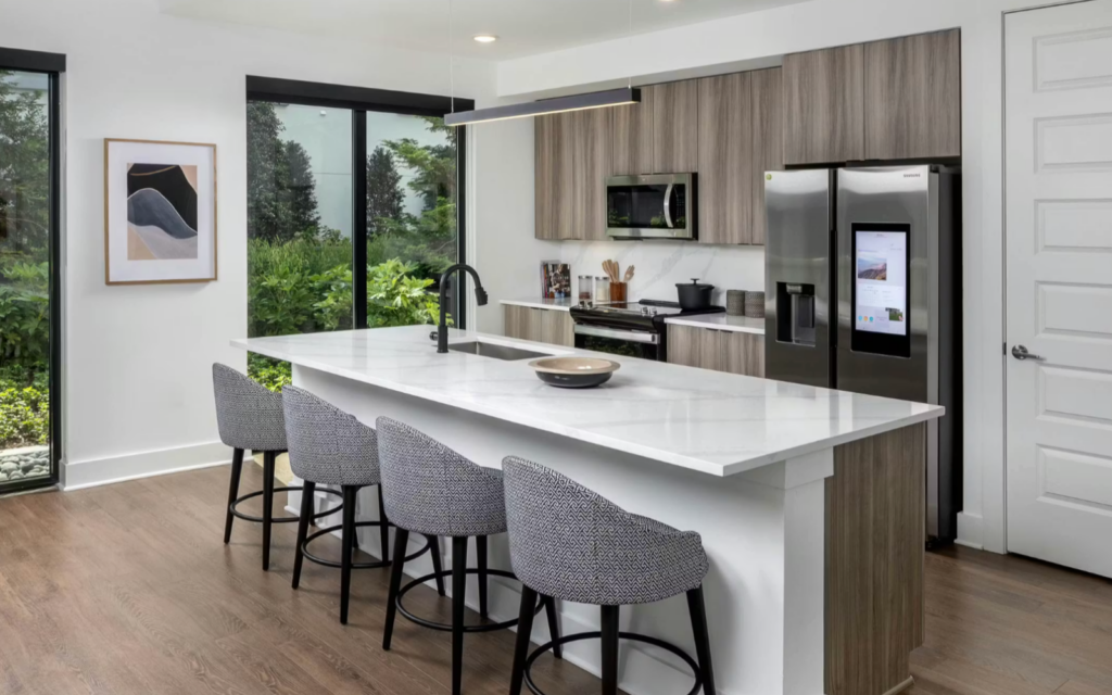 A modern kitchen features a large white marble island with a sleek black faucet and a decorative bowl on top. Behind the island, there are four stylish gray upholstered bar stools with black legs. The cabinetry is a mix of light wood and white, housing stainless steel appliances, including a refrigerator with a touchscreen. Large windows provide a view of lush greenery outside, enhancing the bright and airy atmosphere of the space. A piece of abstract art hangs on the wall, adding a touch of elegance to the decor.