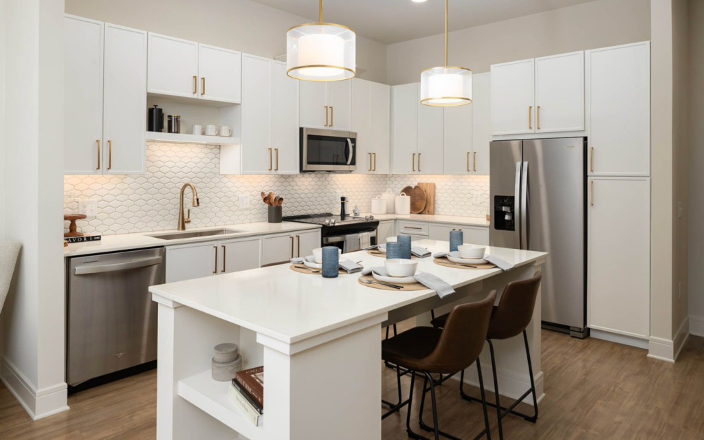 A modern kitchen featuring white cabinetry with gold hardware, a stainless steel refrigerator, and a microwave. The backsplash consists of white hexagonal tiles, and there is a double sink with a gold faucet. A central island with a white countertop is set with blue and white dishware, gray napkins, and woven placemats. Three brown bar stools are positioned at the island, and warm lighting is provided by two pendant lights above. The flooring is a light wood finish, enhancing the contemporary aesthetic.