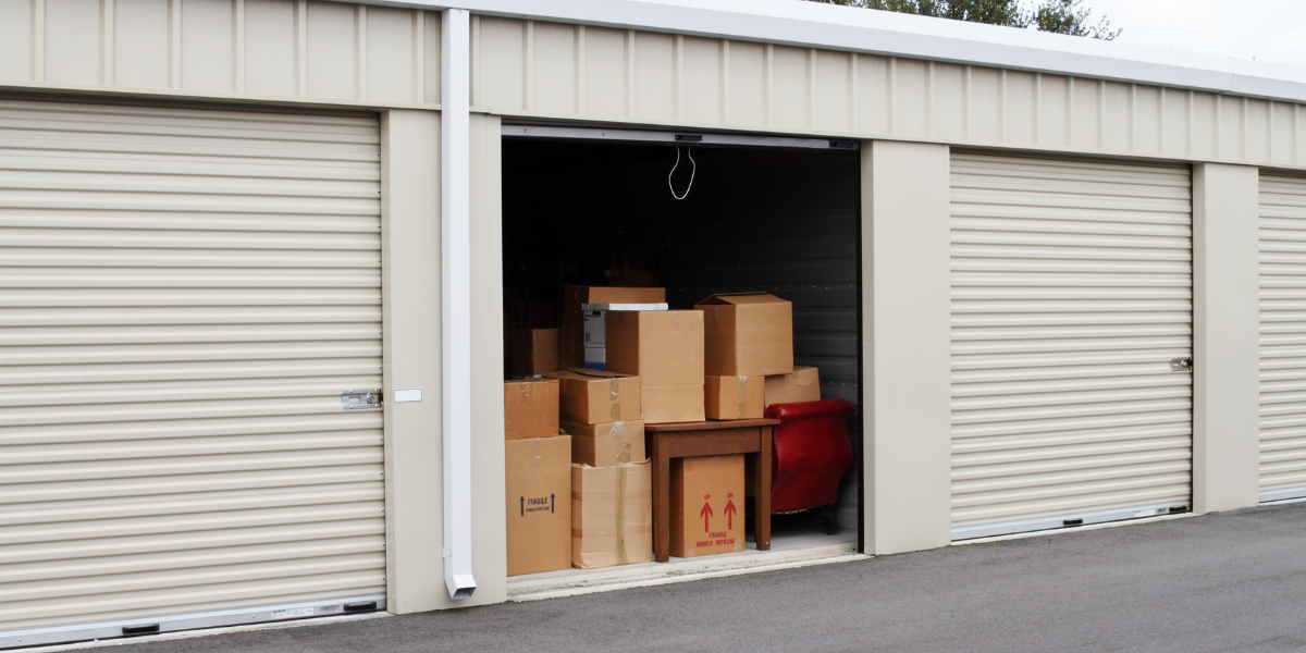 A storage unit with its door partially open reveals a cluttered interior. Inside, there are several cardboard boxes stacked haphazardly, some labeled with "Fragile" warnings. A small wooden table is visible, along with a red upholstered chair. The exterior of the storage unit is beige with a metal roll-up door, and the surrounding area is paved. Sparefoot can help compare storage unit pricing!