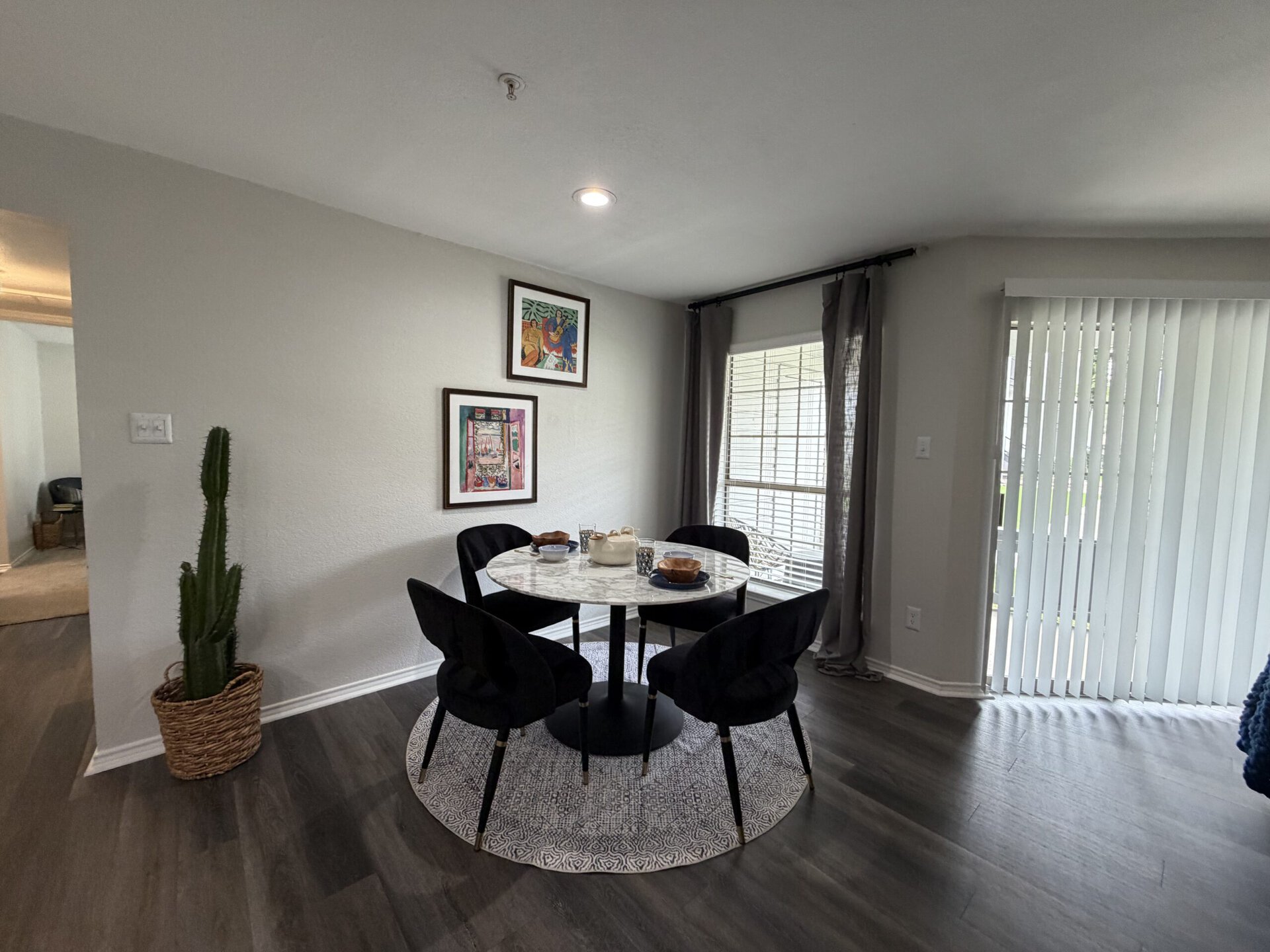 A cozy dining area features a round marble table surrounded by four black upholstered chairs. On the table, there are two wooden bowls, a white ceramic dish, and a few decorative items. A tall cactus in a woven basket stands in the corner, while two framed artworks hang on the wall above the table. Natural light streams in through a window with vertical blinds, complemented by sheer gray curtains. The flooring is a dark wood, enhancing the modern aesthetic of the space.