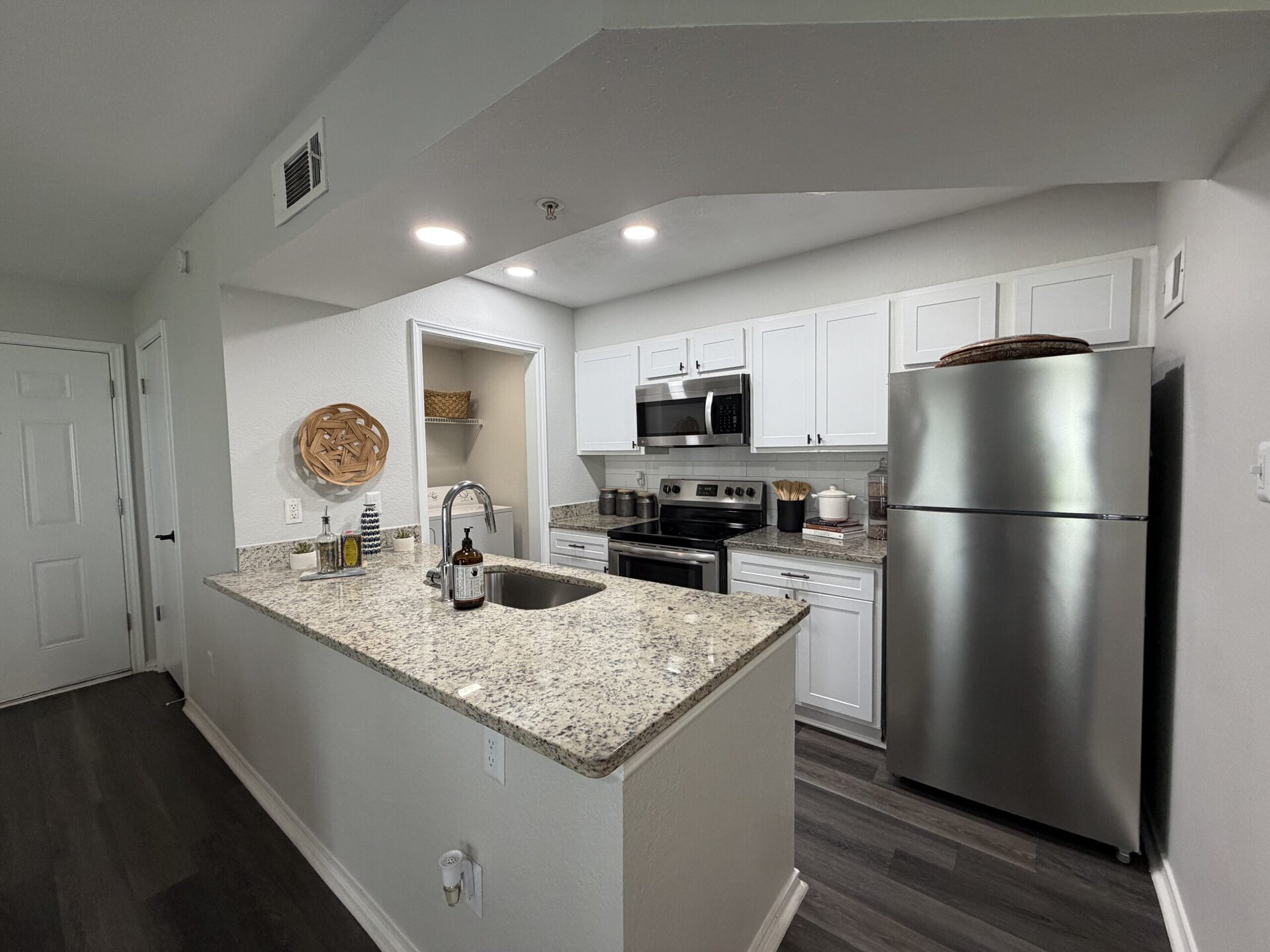 A modern kitchen featuring white cabinetry and granite countertops. The kitchen includes stainless steel appliances, such as a refrigerator, microwave, and oven. A sink with a sleek faucet is positioned on the island, which also has decorative items like a bottle and a small plant. The flooring is dark, contrasting with the light-colored walls. A woven basket is mounted on the wall for added decor, and there is a pantry area visible through an open door. The overall design is clean and contemporary.