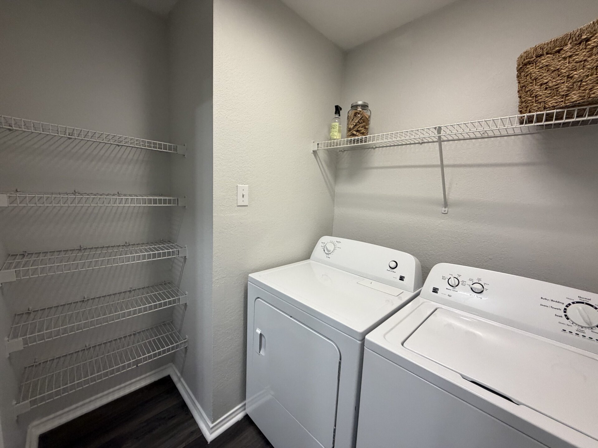 A laundry room features two white appliances: a washing machine and a dryer, positioned side by side. Above them, there are two wire shelves for storage. The walls are painted a light gray, and there is a small jar and a spray bottle on the upper shelf. The floor is dark, contrasting with the light-colored walls and appliances.