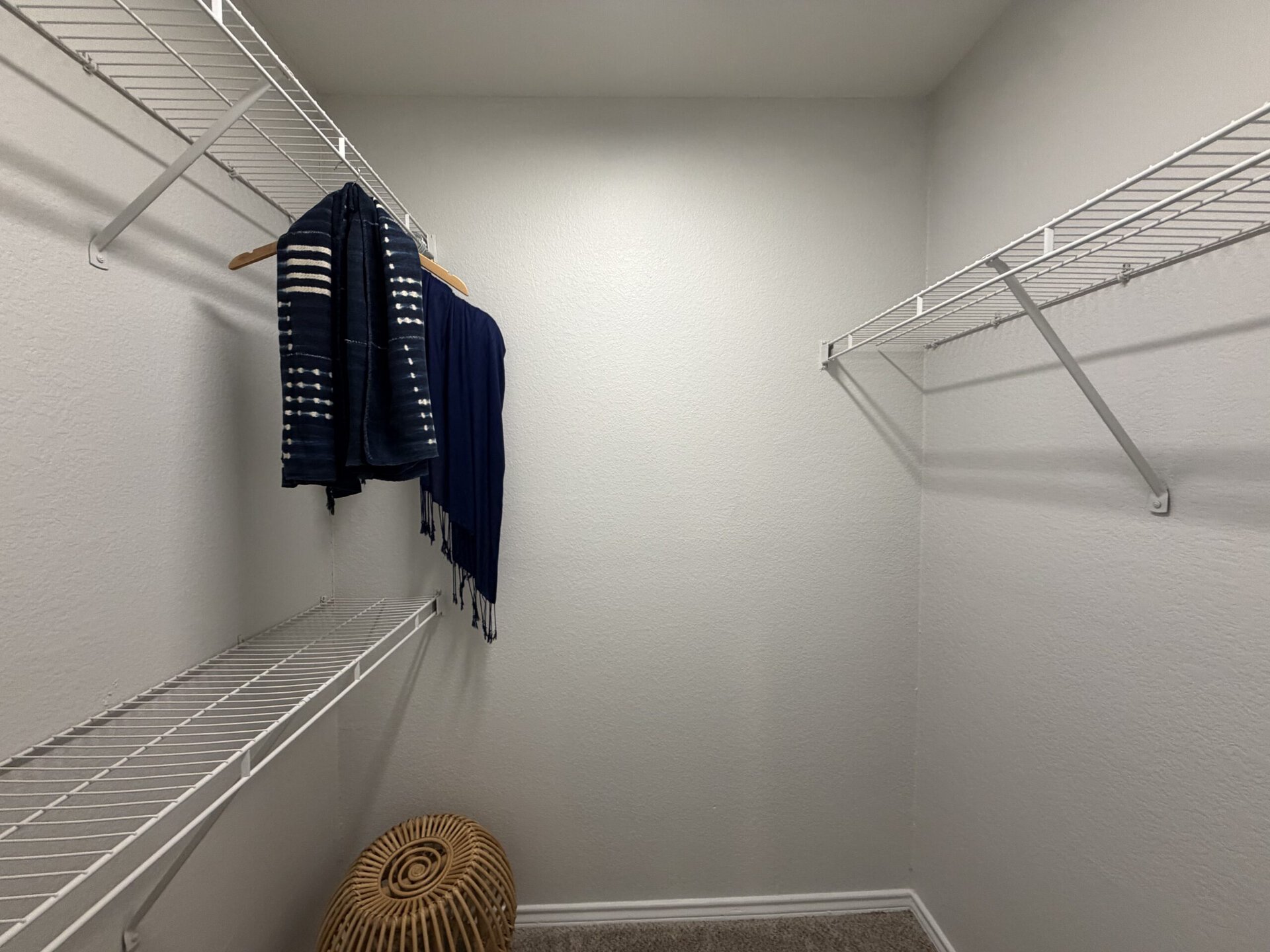 A small, empty closet features two wire shelves and a hanging rod. On the rod, there are two dark blue garments, one with a patterned design. The walls are painted a light color, and the floor is bare. The overall space appears tidy and organized.