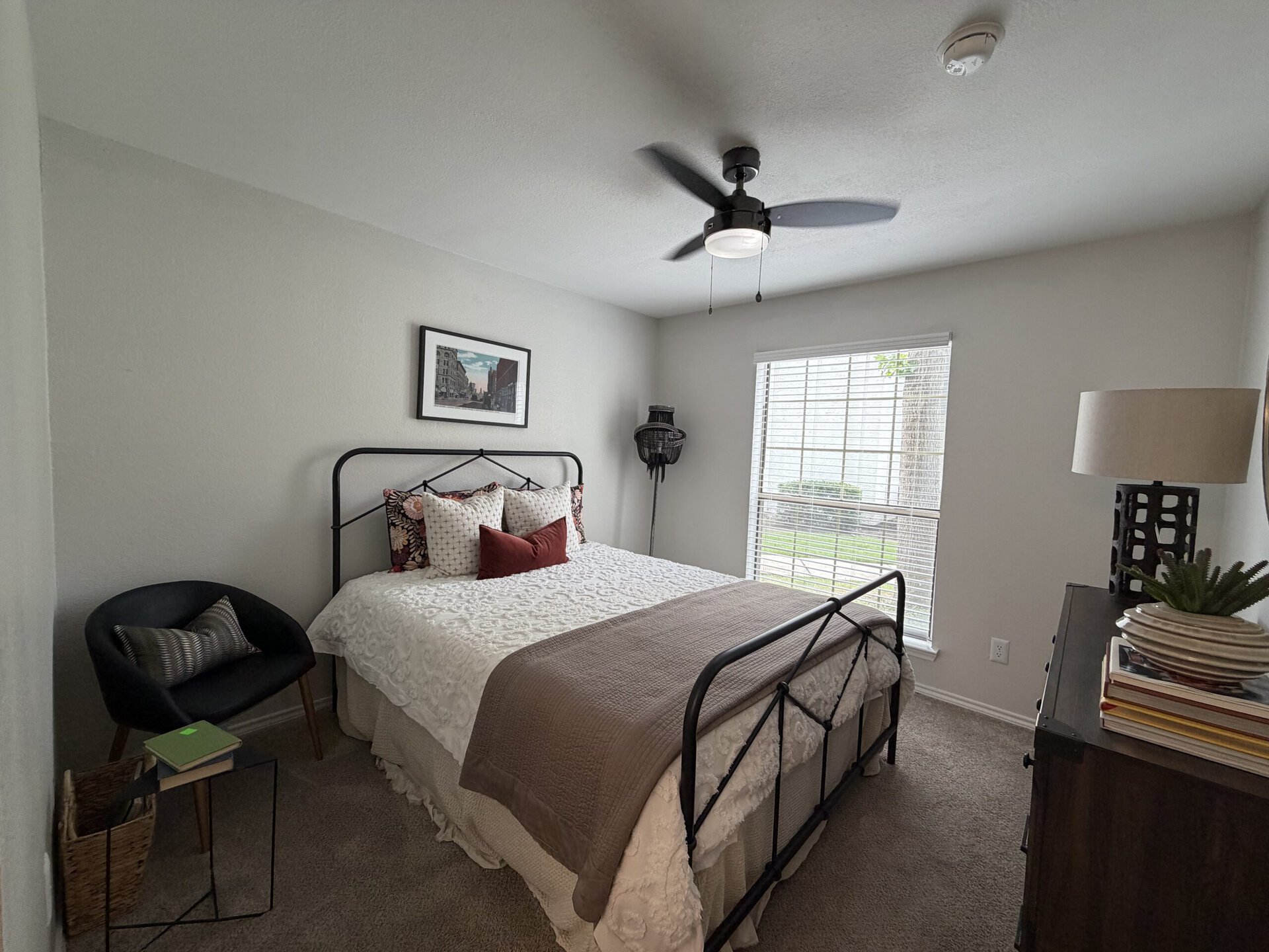 A cozy bedroom features a black metal bed frame with a white quilted bedspread and decorative pillows in various patterns, including floral and solid colors. A bedside table holds a stack of books and a small plant. A black accent chair with a patterned cushion is positioned nearby. Natural light streams in through a window with blinds, illuminating the room's neutral walls and carpet. A ceiling fan adds a modern touch, and a framed artwork hangs above the bed, enhancing the decor.