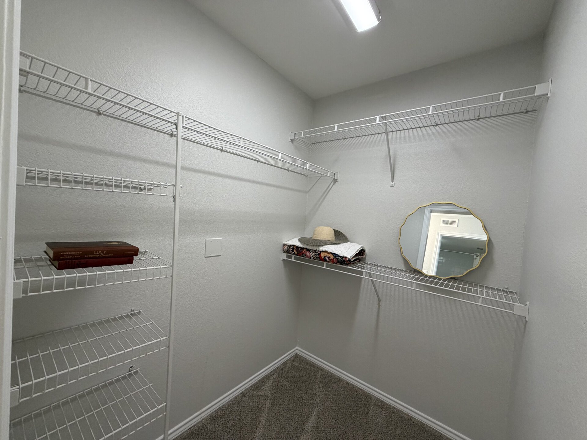 A small closet features white wire shelving on three walls. On one shelf, there are two stacked books and a folded towel with a patterned design. A wide-brimmed hat rests on top of the towel. A round mirror with a decorative gold frame is mounted on the wall, reflecting the light. The walls are painted a light color, and the floor is carpeted.
