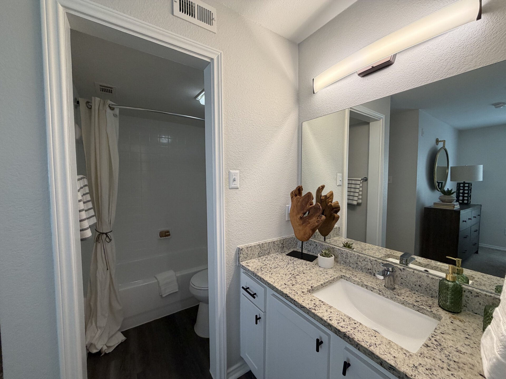 A modern bathroom features a light-colored wall and a granite countertop with a white sink. A decorative wooden sculpture stands on the counter alongside a small potted plant and a green soap dispenser. To the left, a shower area is partially visible, with a white curtain and a towel hanging nearby. A mirror above the sink reflects the space, while a round mirror and a lamp are seen in the background, adding to the room's decor.