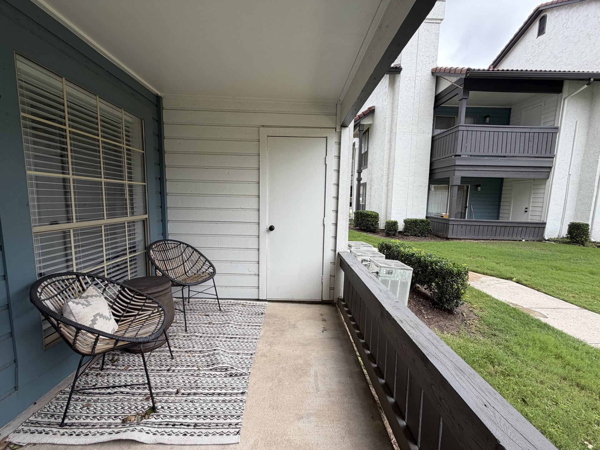 A small porch area features two black wicker chairs with a decorative pillow and a round wooden table. The floor is covered with a patterned rug. A white door is visible on one side, leading to an interior space. In the background, there are two neighboring buildings with balconies, and neatly trimmed bushes line the grassy area. The overall atmosphere is inviting and cozy.