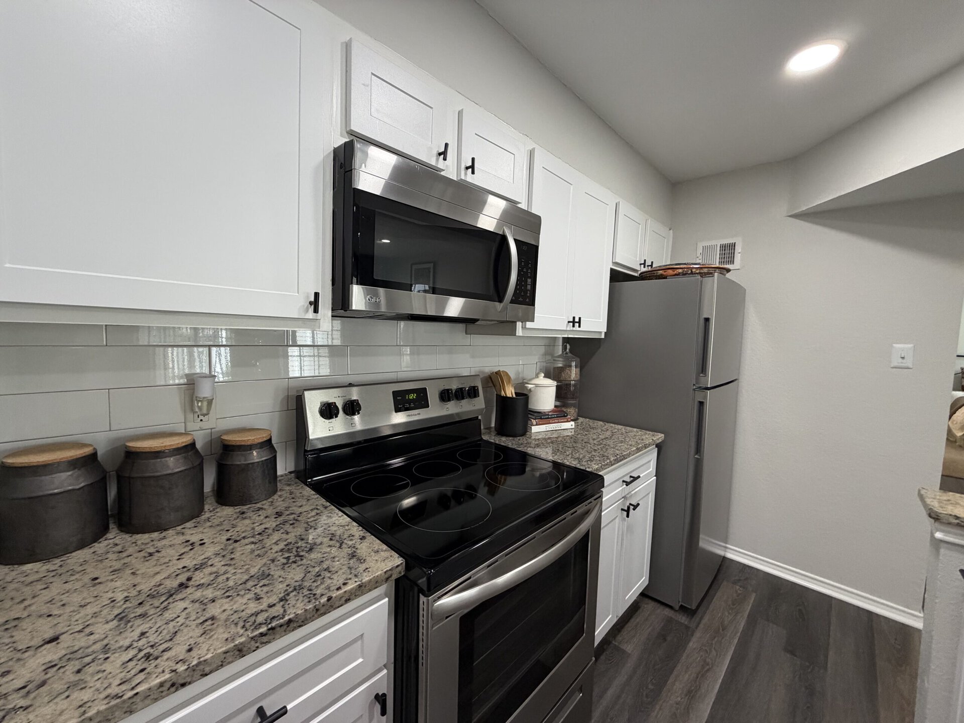 A modern kitchen features white cabinetry with black hardware, a stainless steel microwave mounted above a black electric stove. The countertop is made of speckled granite, and there are three dark containers with wooden lids arranged neatly. A silver refrigerator stands against the wall, and a few cookbooks are stacked beside a white ceramic container holding wooden utensils. The backsplash consists of light gray subway tiles, enhancing the contemporary look of the space.