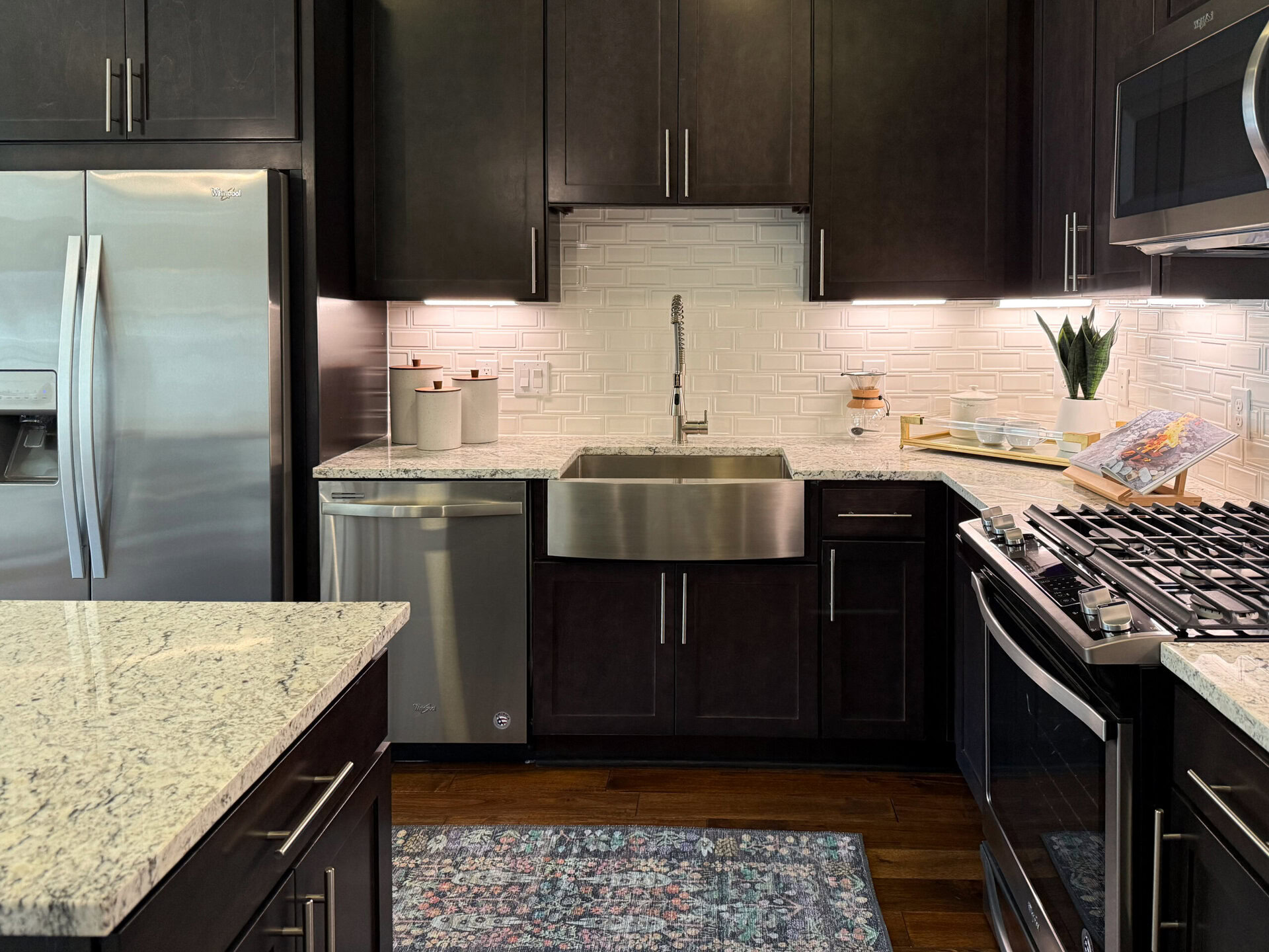 A modern kitchen features dark cabinetry and a large stainless steel farmhouse sink. The countertop is made of light-colored granite, contrasting with the dark wood. To the left, there is a stainless steel refrigerator and a dishwasher. Above the sink, a stylish faucet is mounted against a white tiled backsplash with a textured pattern. On the countertop, there are several decorative containers, a small plant, and a wooden tray holding glass bowls and a cookbook displayed on a stand. The overall design is sleek and contemporary, with warm lighting enhancing the space.