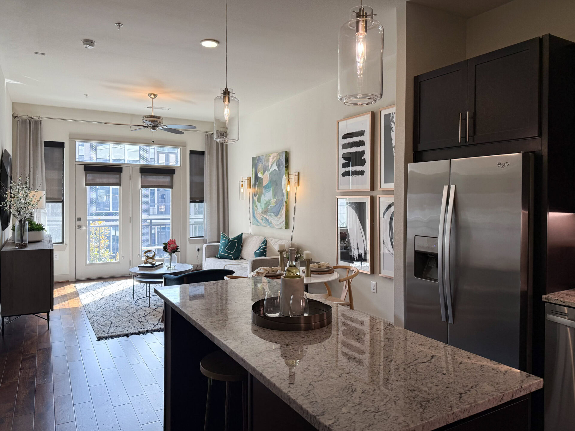 A modern kitchen and living area are depicted, featuring a sleek granite countertop with a dark base. In the foreground, a tray holds glasses and a bottle. The kitchen includes stainless steel appliances, including a refrigerator. The living area showcases a light-colored sofa adorned with teal cushions, a round coffee table, and a decorative rug. Large windows allow natural light to fill the space, highlighting wall art and stylish lighting fixtures. A potted plant adds a touch of greenery to the decor.