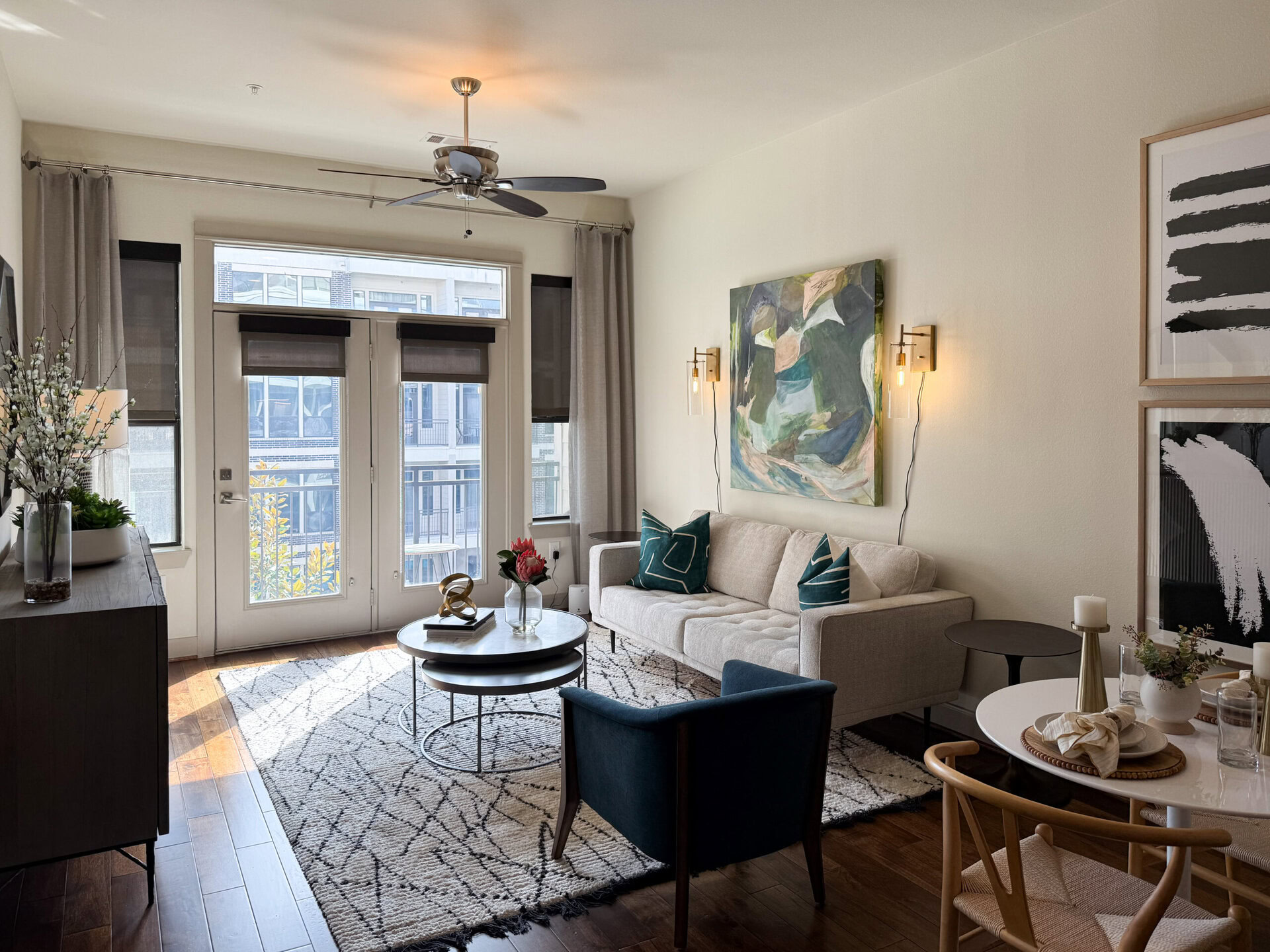 A cozy living room features a light-colored sofa adorned with teal and white patterned cushions. A round coffee table sits in front, accompanied by a small side table. The space is illuminated by a wall-mounted light fixture and natural light streaming through large windows with blinds. A piece of abstract art hangs on the wall, while decorative elements like a vase with flowers and a small plant add a touch of greenery. The floor is covered with a patterned rug, enhancing the inviting atmosphere.