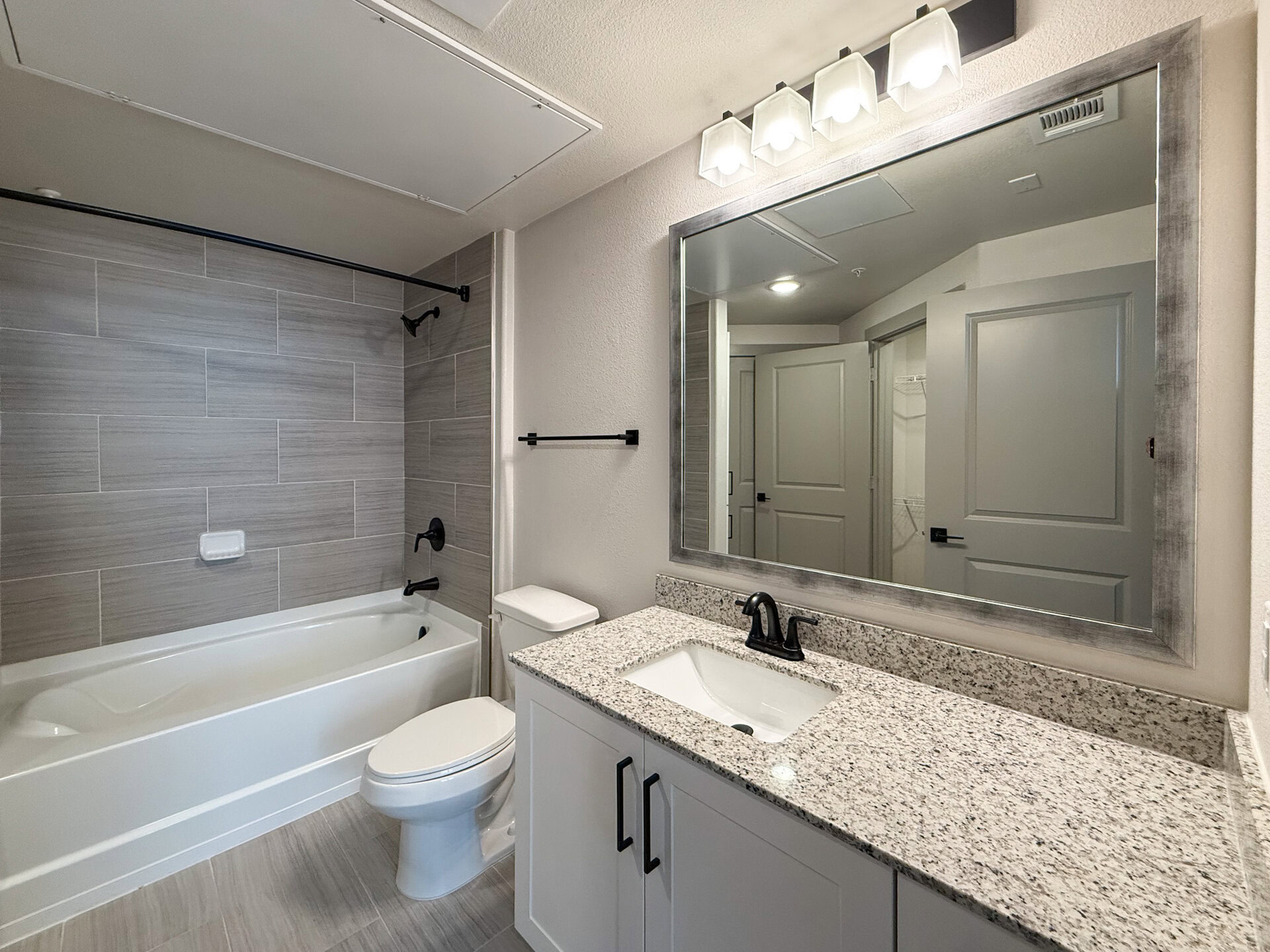 A modern San Antonio apartment bathroom features a white bathtub with a black shower rod and faucet. The walls are adorned with light gray tiles, creating a sleek look. A white toilet is positioned next to a granite countertop with a rectangular sink and a black faucet. Above the sink, a large mirror with a silver frame reflects the space, which includes a door leading to a closet area. The overall design is clean and contemporary, with neutral tones and stylish fixtures.