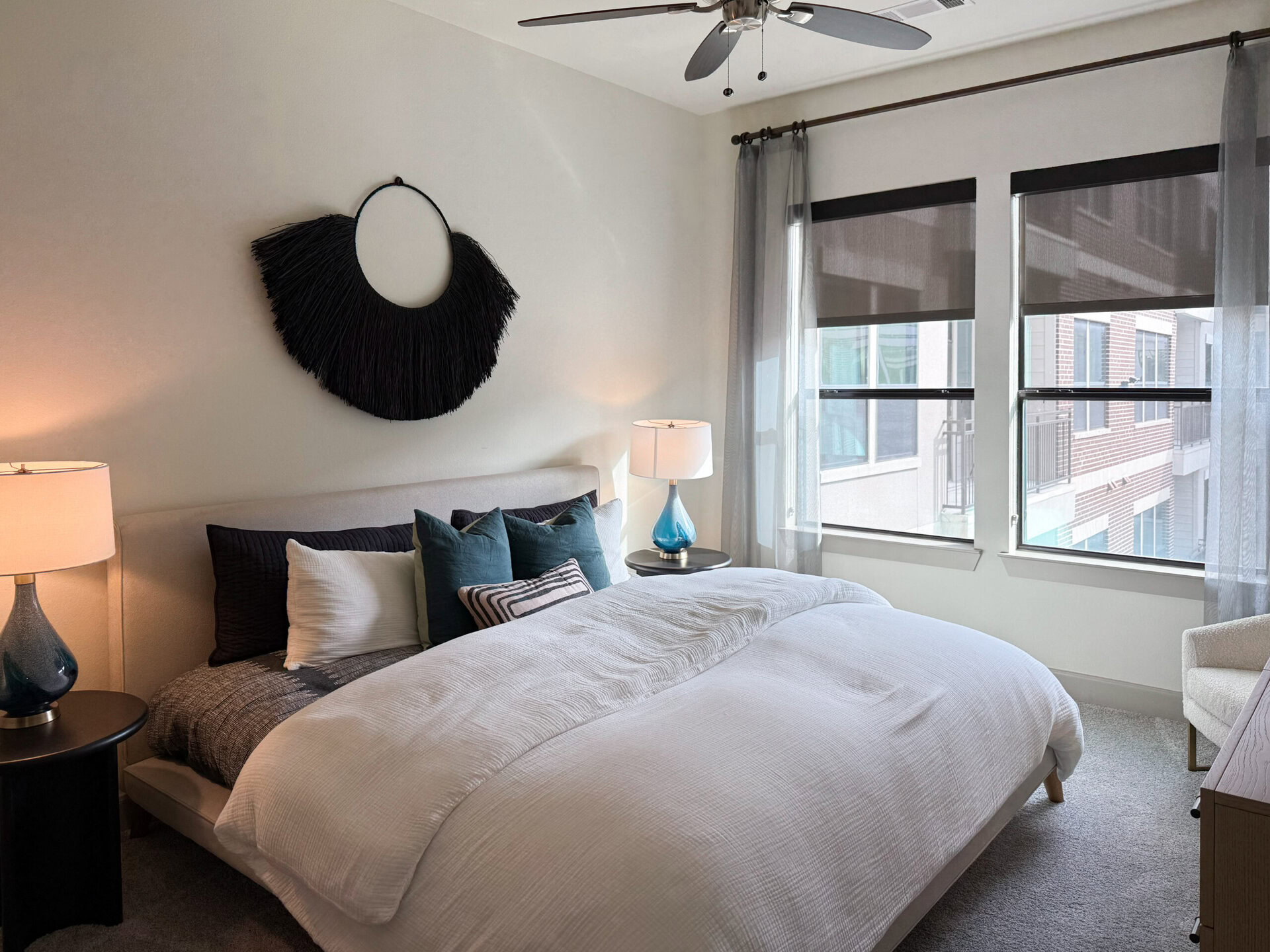 A modern bedroom features a large bed with a white comforter and an assortment of decorative pillows in shades of blue and black. Above the bed, a circular black wall hanging adds a stylish touch. On one side of the bed, a bedside table holds a lamp with a blue base and a white shade. Two large windows with sheer curtains allow natural light to enter, revealing a view of neighboring buildings. A cozy armchair sits in the corner, complementing the overall contemporary design.