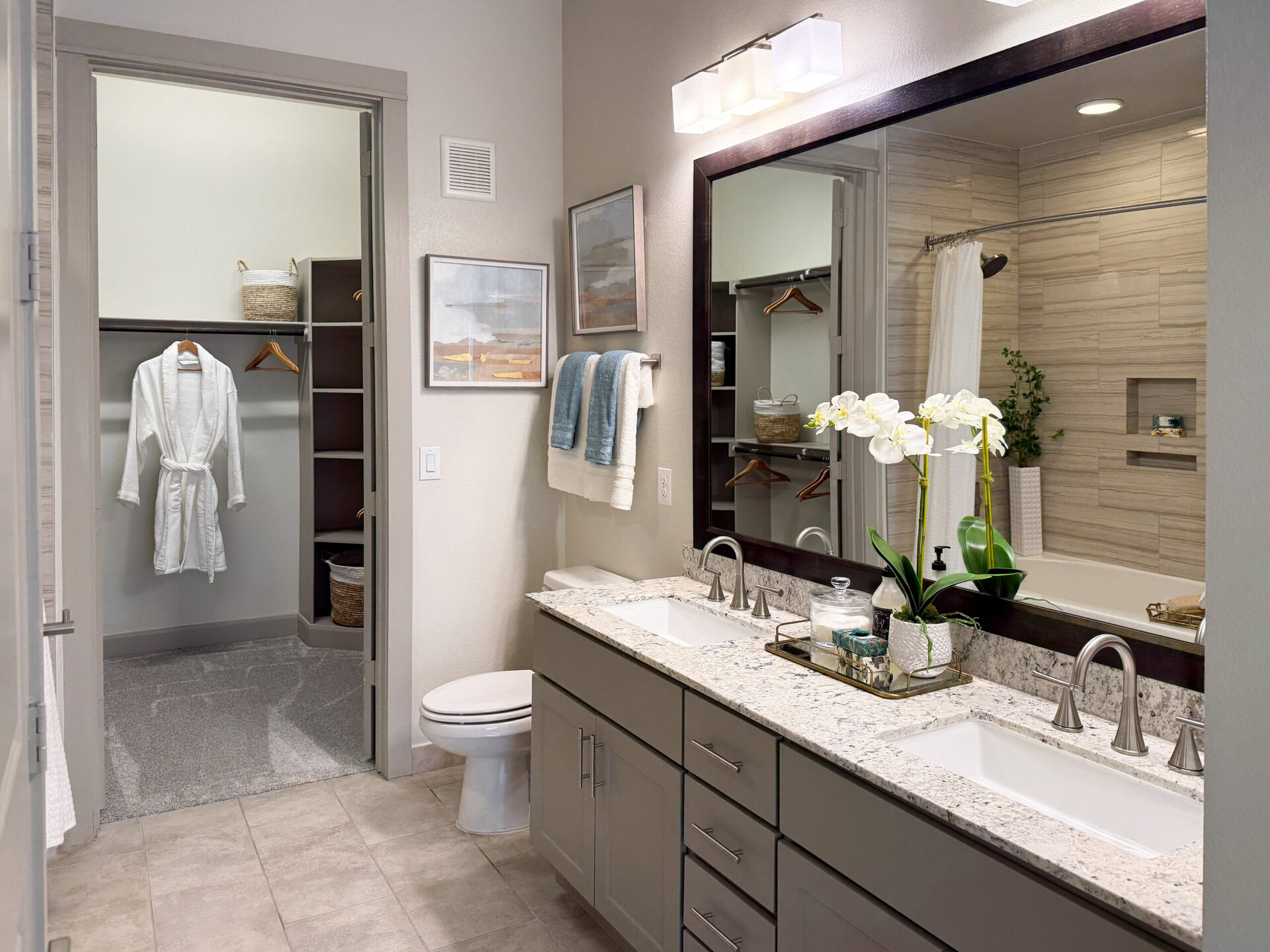 A modern bathroom features a double vanity with a granite countertop and two sinks. Above the vanity, a large mirror reflects the space. On the countertop, there are decorative items including a glass container and potted plants. A white robe hangs in an open closet to the left, which has shelves and baskets for storage. The bathroom includes a toilet and a bathtub with a shower curtain. Soft lighting and neutral colors create a calming atmosphere.