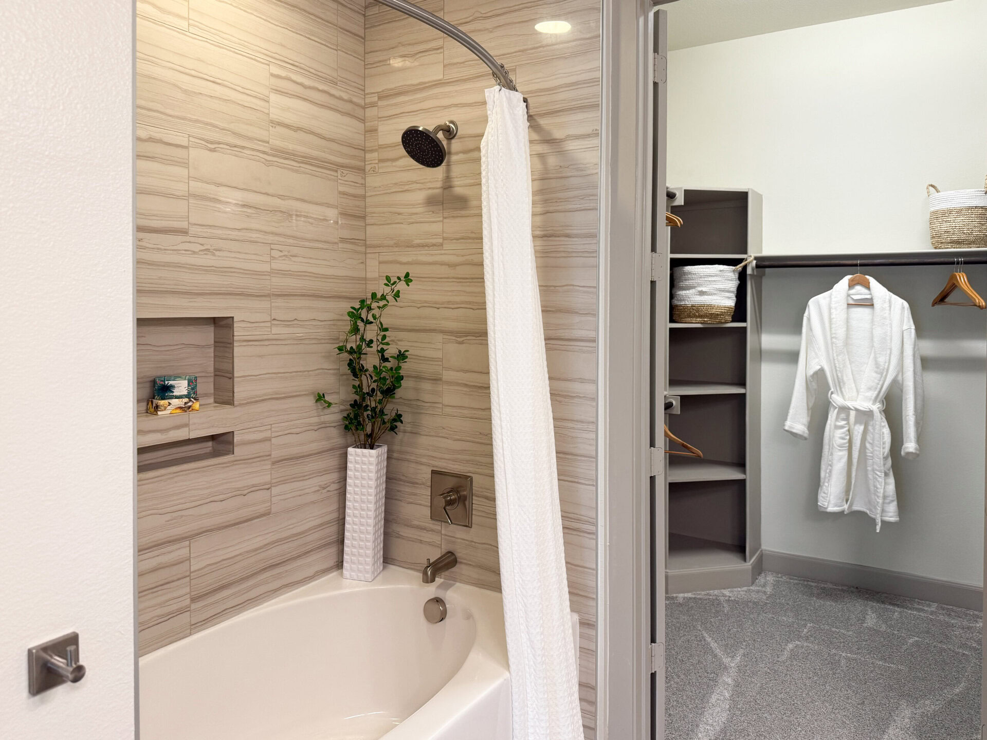 A modern bathroom features a shower area with a rainfall showerhead and a white curtain. The walls are adorned with light-colored, textured tiles. A small recessed shelf holds decorative items, including a box of soaps. A tall, white vase with greenery adds a touch of nature. Adjacent to the shower is a closet space with neatly organized shelves, displaying rolled towels and woven baskets. A plush white robe hangs on a wooden hanger, completing the serene and stylish atmosphere.