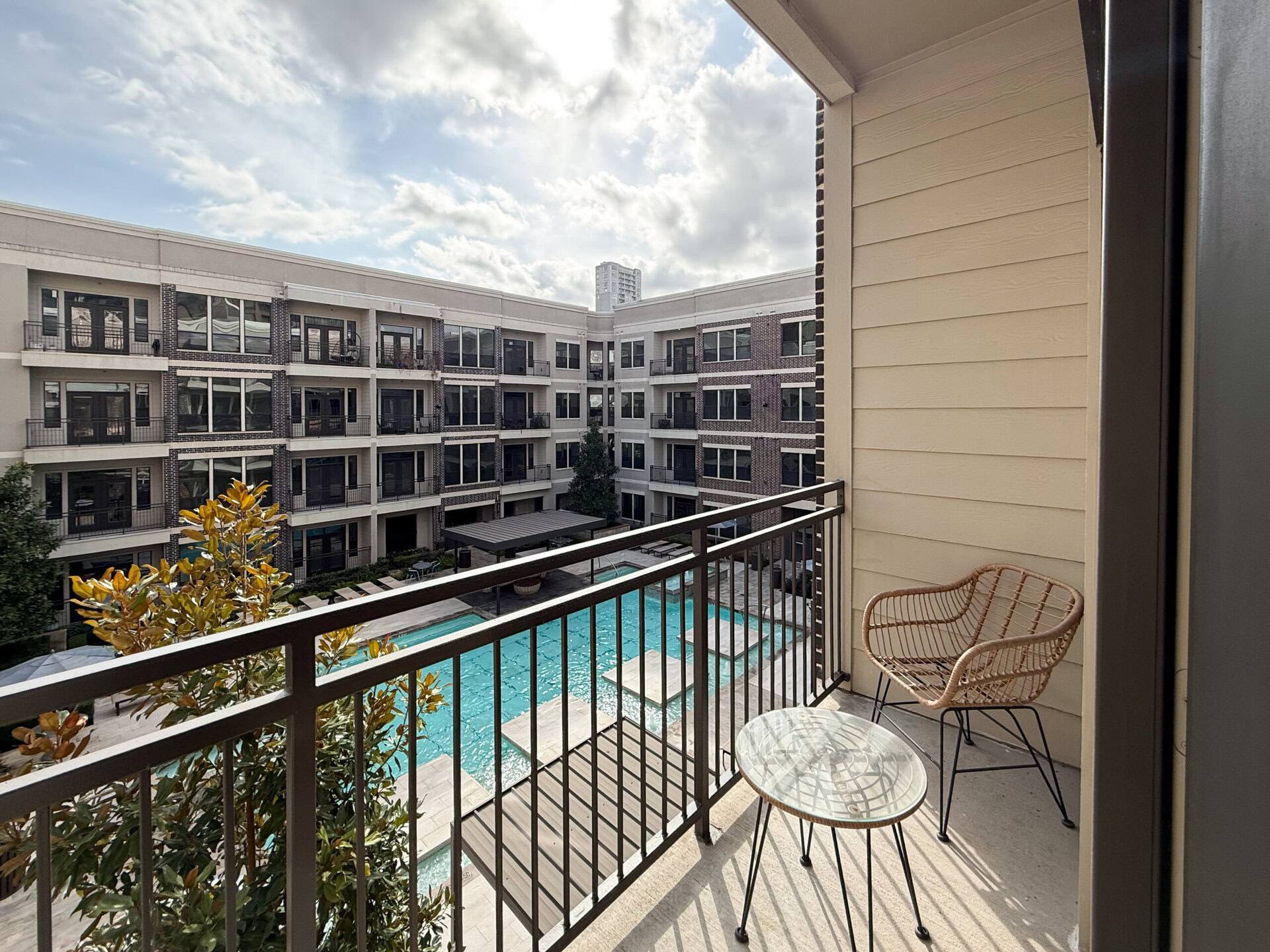 A balcony overlooks a modern apartment complex courtyard featuring a swimming pool. The balcony has a round glass table and a woven chair. In the courtyard, there are several lounge chairs and a shaded seating area with a pergola. Surrounding the pool are landscaped areas with greenery and decorative plants. The sky is partly cloudy, adding a soft light to the scene.