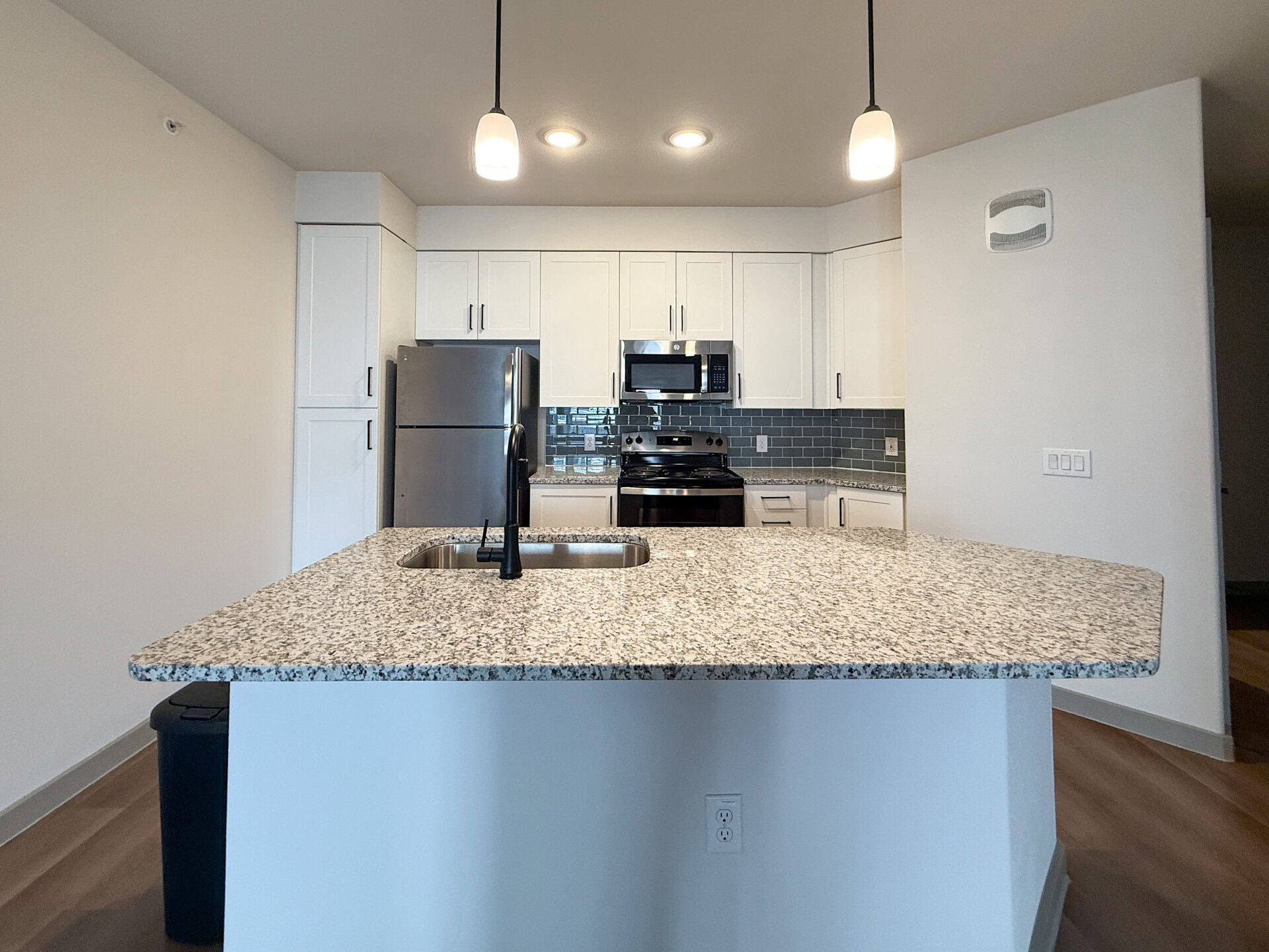 A modern San Antonio apartment kitchen featuring a large granite countertop with a sink and a sleek black faucet. The cabinetry is white with simple handles, and stainless steel appliances, including a refrigerator and an oven, are visible. A backsplash of blue-green tiles adds a pop of color behind the stove and microwave. The lighting is provided by pendant lights above the countertop, creating a bright and inviting atmosphere. The flooring is light wood, enhancing the contemporary feel of the space.