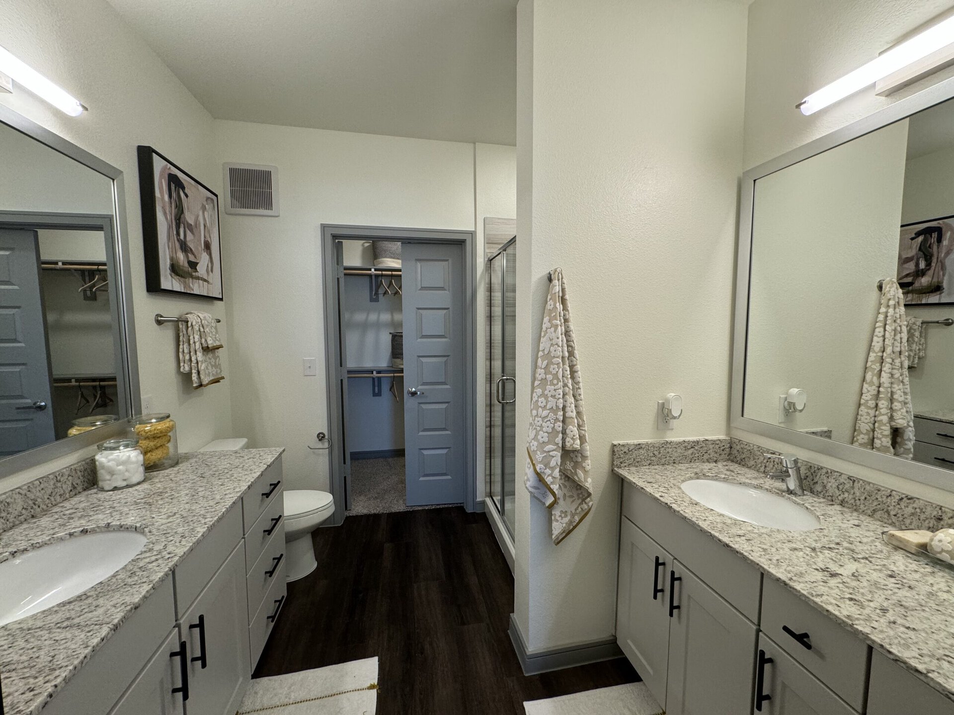 A modern bathroom features a double vanity with a light gray granite countertop and two sinks. Above the vanity, there are two large mirrors. A decorative piece hangs on the wall, adding a touch of art. To the left, a door leads to a closet with open shelving. A glass shower enclosure is visible next to the vanity, and a toilet is positioned nearby. The flooring is dark, contrasting with the light walls and cabinetry. A towel hangs on a rack, and decorative jars with cotton balls and bath items are placed on the countertop.