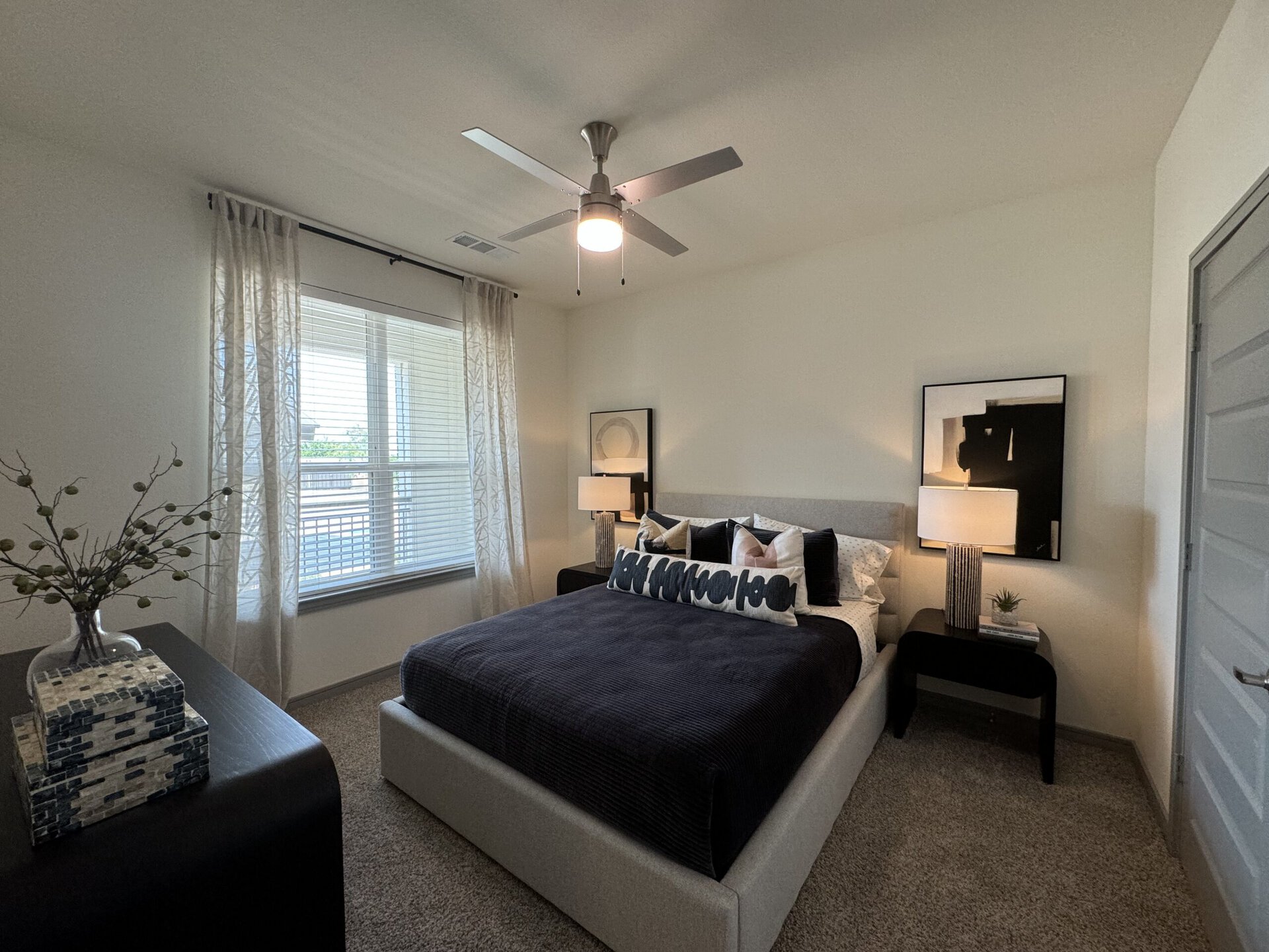 A cozy bedroom features a large bed with a dark, textured blanket and an array of decorative pillows. A bedside table holds a modern lamp and a small plant, while a decorative vase with branches adds a touch of greenery. Natural light streams in through a window adorned with sheer curtains, and a piece of abstract art hangs on the wall above the bed. The room has a neutral color palette with soft carpeting and a contemporary ceiling fan.