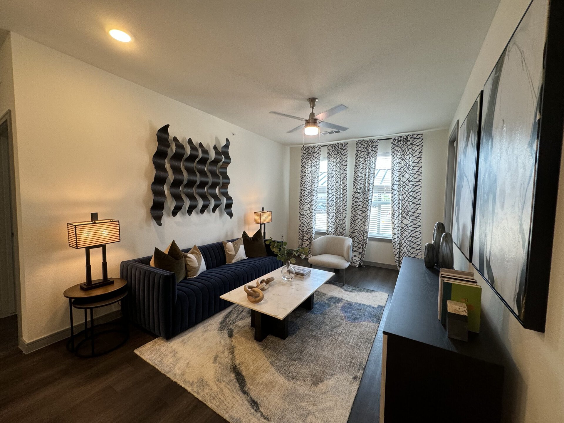 A modern living room features a navy blue, tufted sofa adorned with various decorative pillows in shades of green and white. A round side table with a black base holds a stylish lamp with a striped shade, providing warm light. In front of the sofa, a rectangular marble coffee table displays a decorative wooden piece and a small vase with greenery. The room is brightened by large windows dressed with patterned curtains. On the wall, a wavy black art piece adds texture, while a dark console table displays decorative items and books. The flooring is a dark wood, complementing the overall contemporary aesthetic.