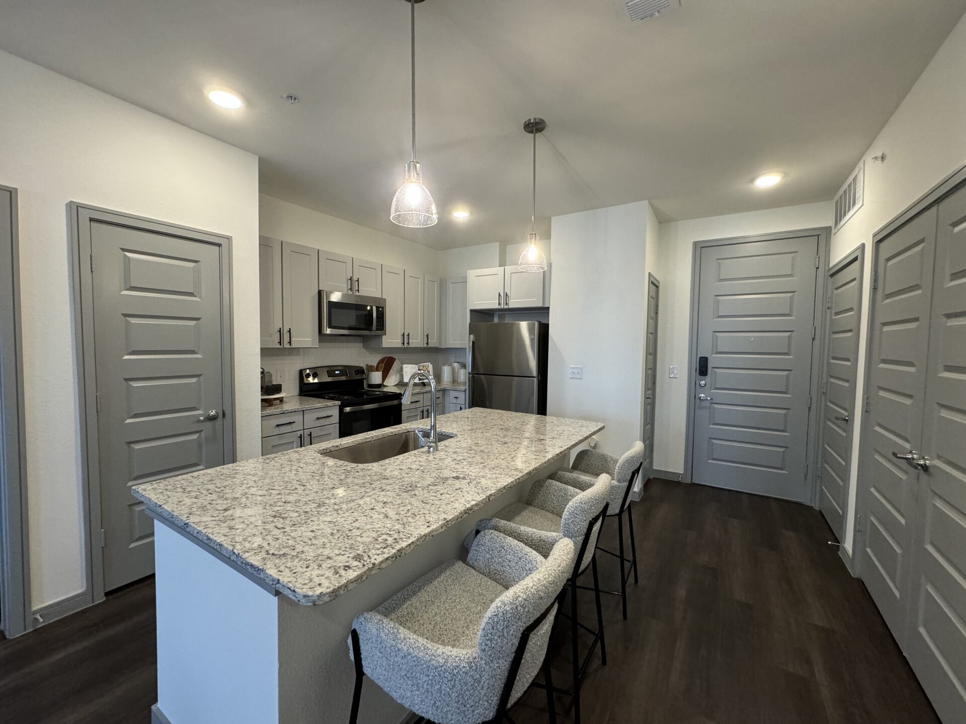 A modern kitchen features light gray cabinetry and stainless steel appliances, including a microwave and refrigerator. The countertop is made of speckled granite, with a sink integrated into the island. Three upholstered bar stools with black legs are positioned at the island. The space is well-lit with pendant lights hanging above. To the right, several closed doors suggest additional rooms or storage, and the flooring is dark wood, adding contrast to the lighter elements of the kitchen.