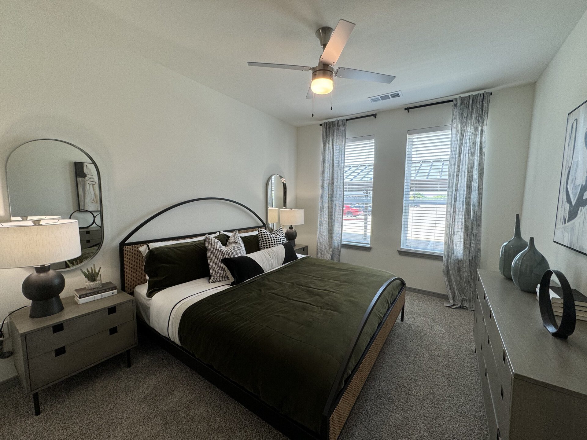 A modern bedroom features a large bed with a dark green velvet comforter and decorative pillows in various patterns. Beside the bed, a nightstand holds a black lamp with a round base and a small plant. A round mirror hangs above the nightstand, reflecting light. Two windows with sheer curtains allow natural light to enter, and a dresser with decorative vases and books is positioned against the wall. The room has a neutral color palette and a cozy carpeted floor.